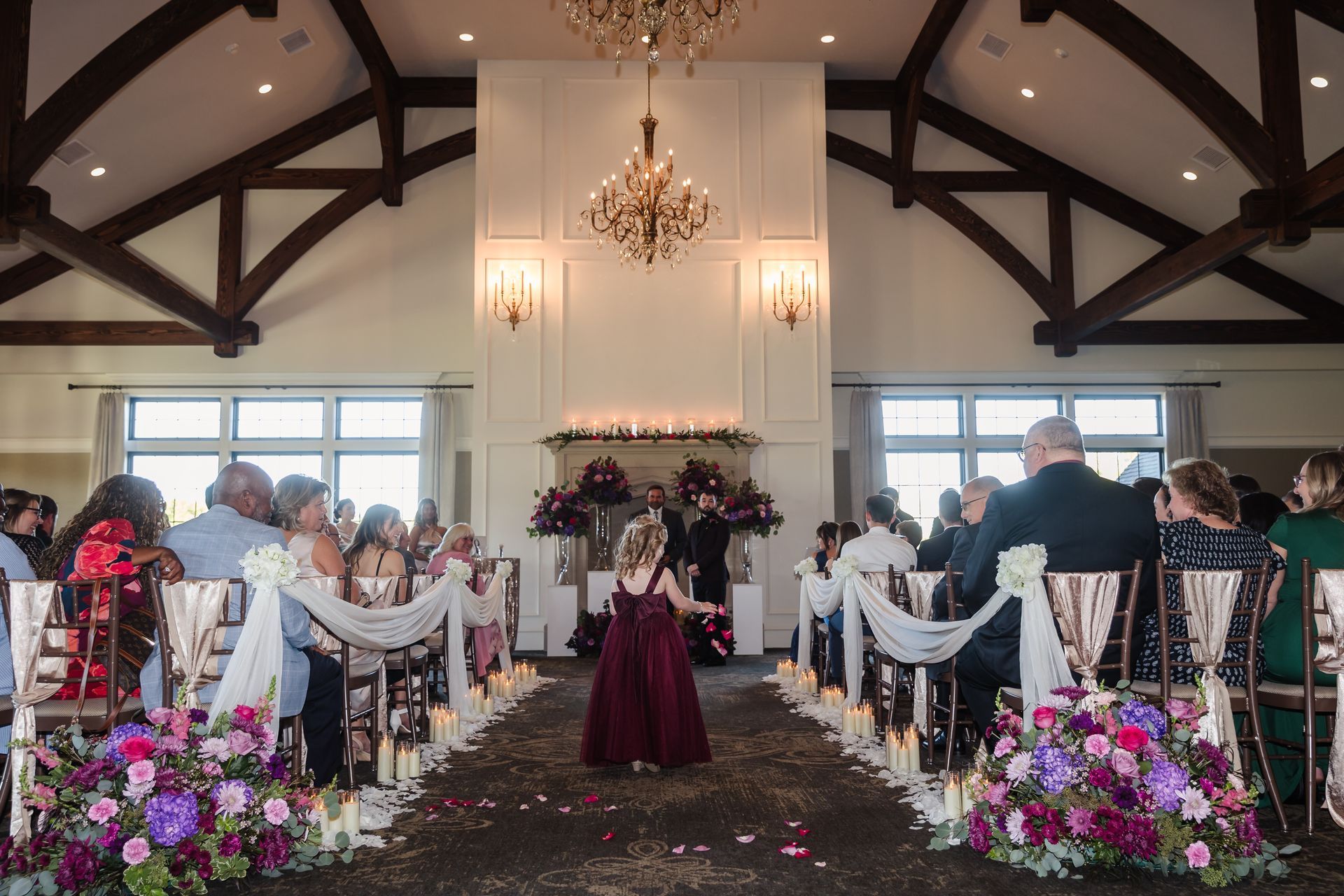 Wedding ceremony in a hall with flower aisle, chandelier, and guests seated.