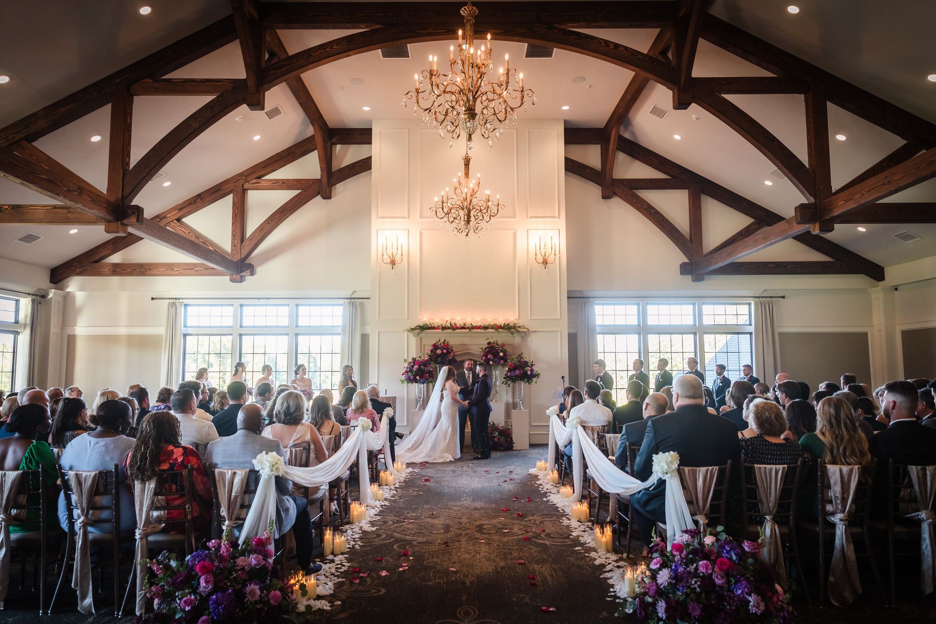Wedding ceremony in a large room with high wooden beams and chandeliers. Bride and groom at the altar, guests seated.