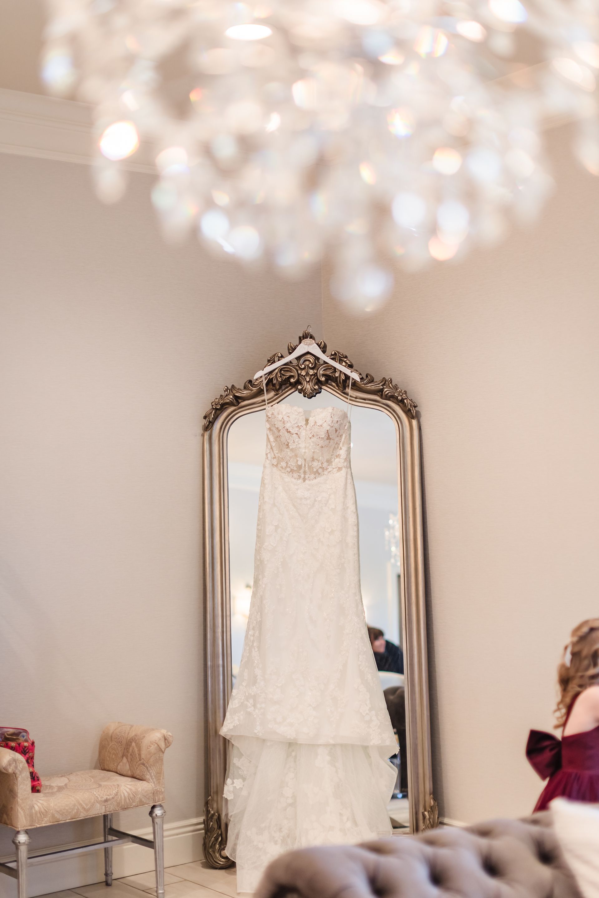 Wedding dress hanging in front of a mirror, with a chandelier above.