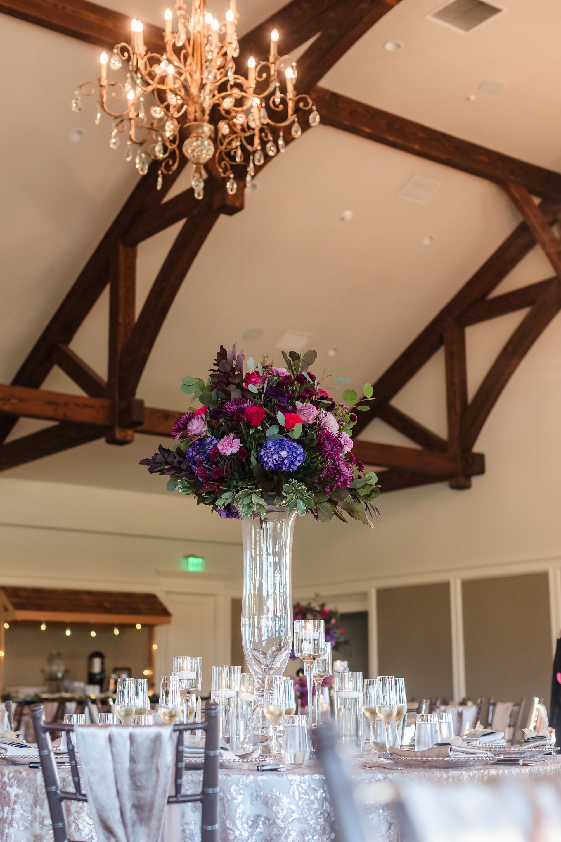 Wedding reception table with large floral centerpiece, chandelier, and wooden beams.