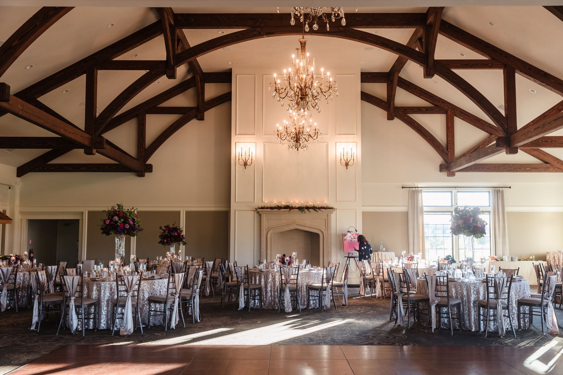 Elegant ballroom set for a wedding reception. Tables with floral centerpieces, chandelier, exposed wooden beams.