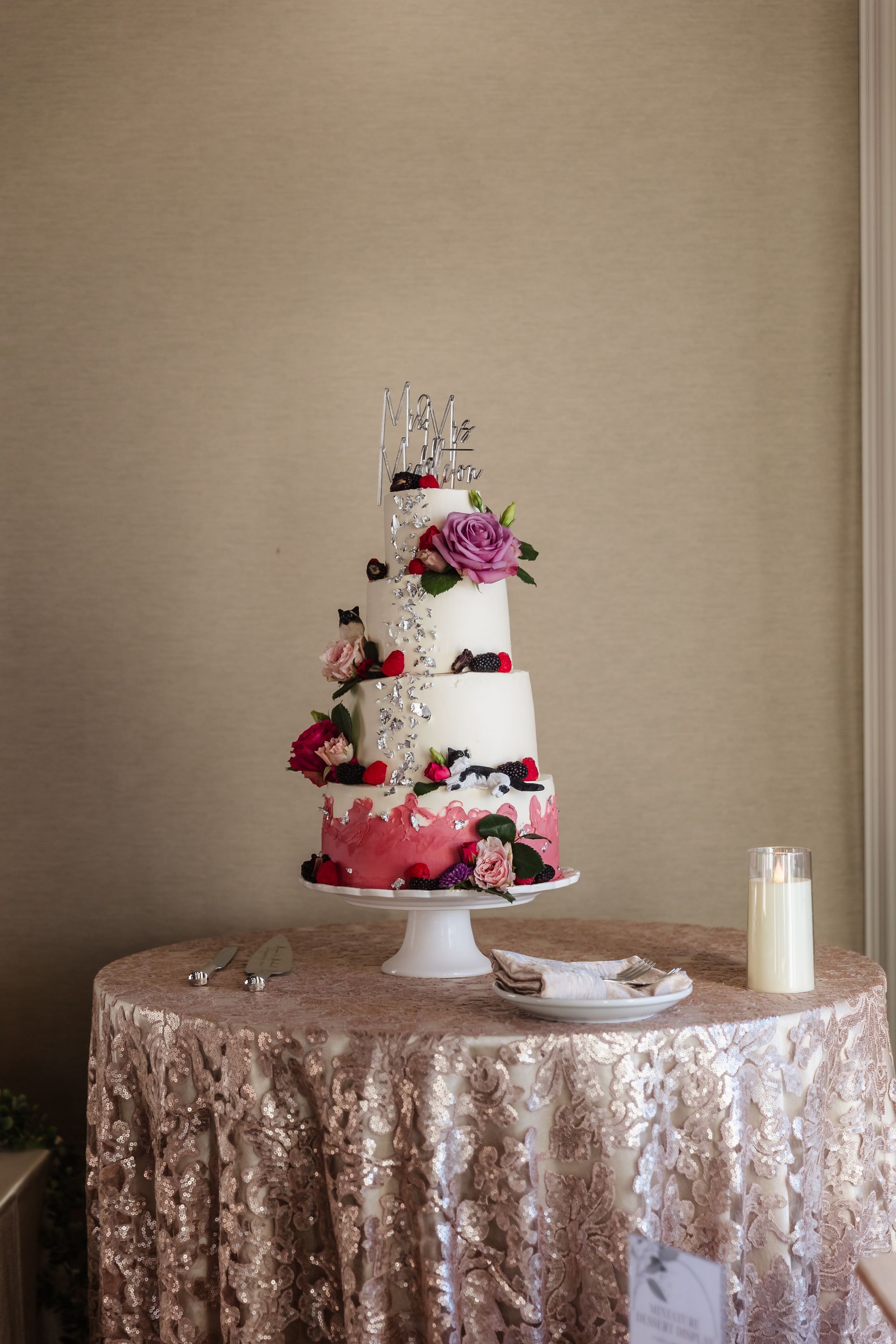 Four-tiered wedding cake, decorated with flowers, berries, and pink frosting, on a sparkly gold table.