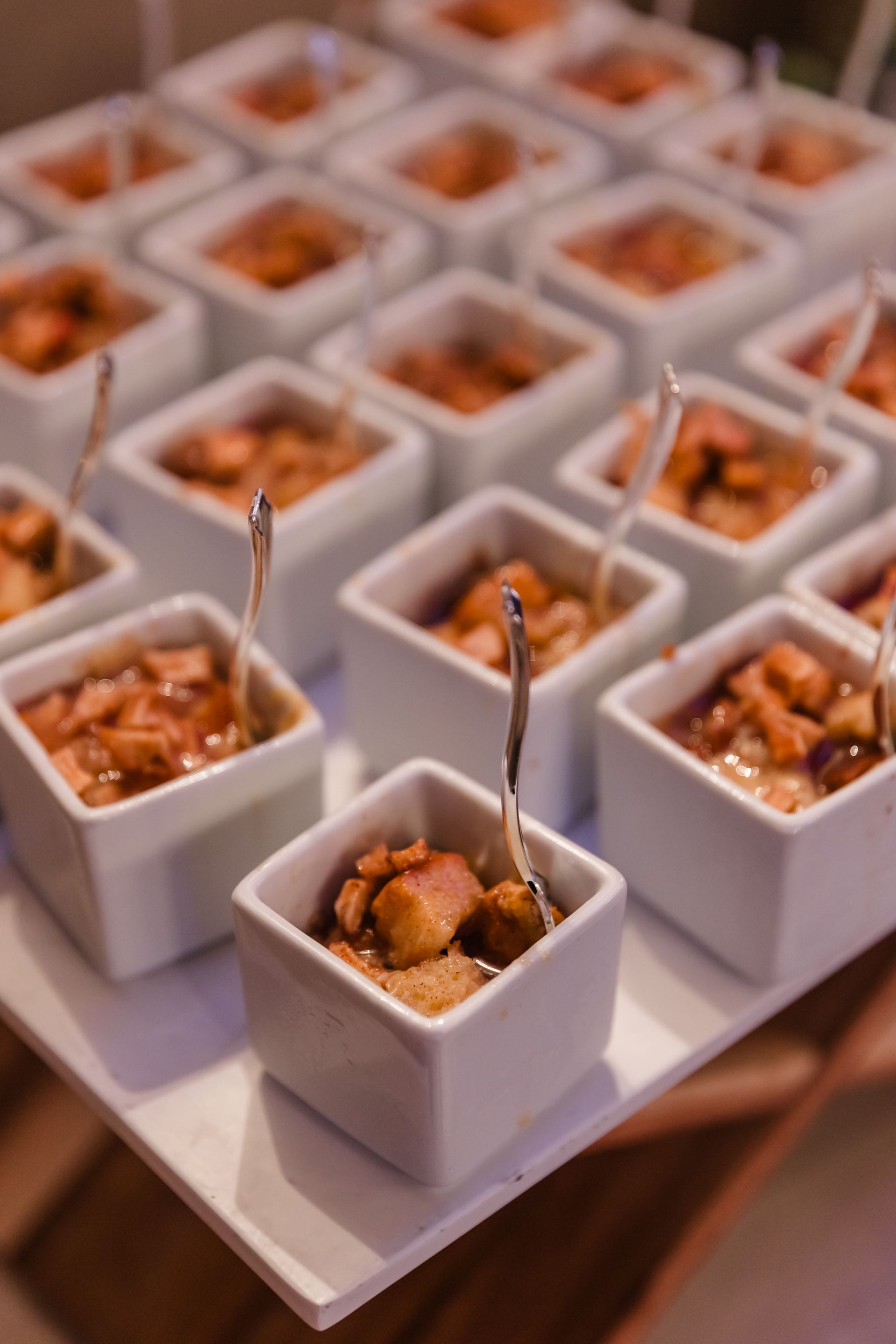 Small white square dishes filled with food, each with a small spoon, arranged on a white tray.