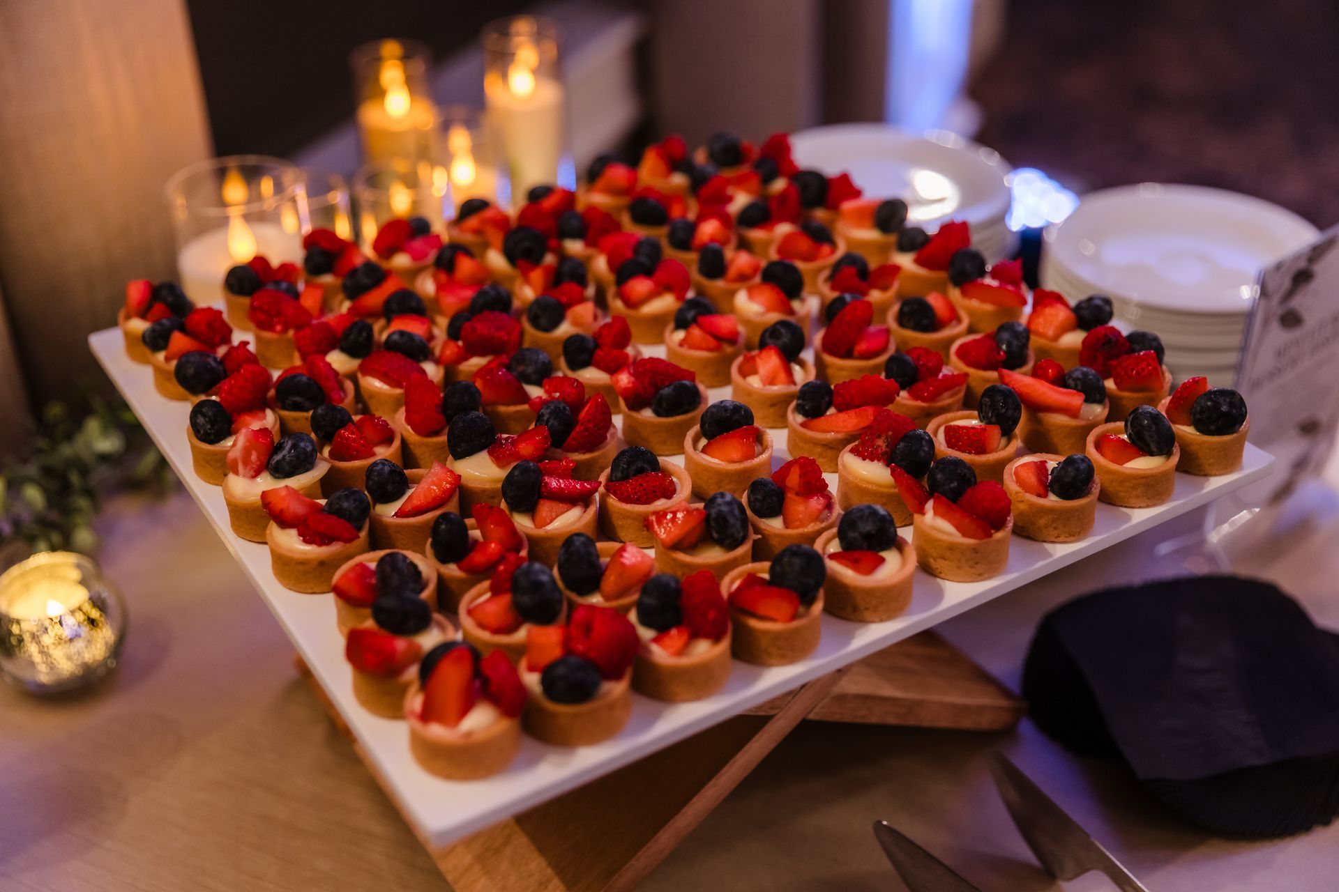 Tray of mini fruit tarts with strawberries, blueberries, blackberries, and cream on a table with candles.
