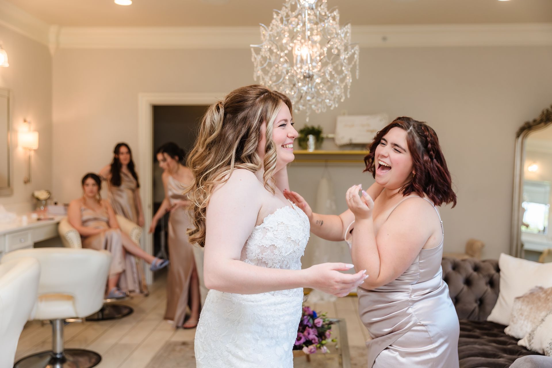 Bride and bridesmaid laugh in a bridal suite. Bridesmaids in gold dresses stand or sit nearby.