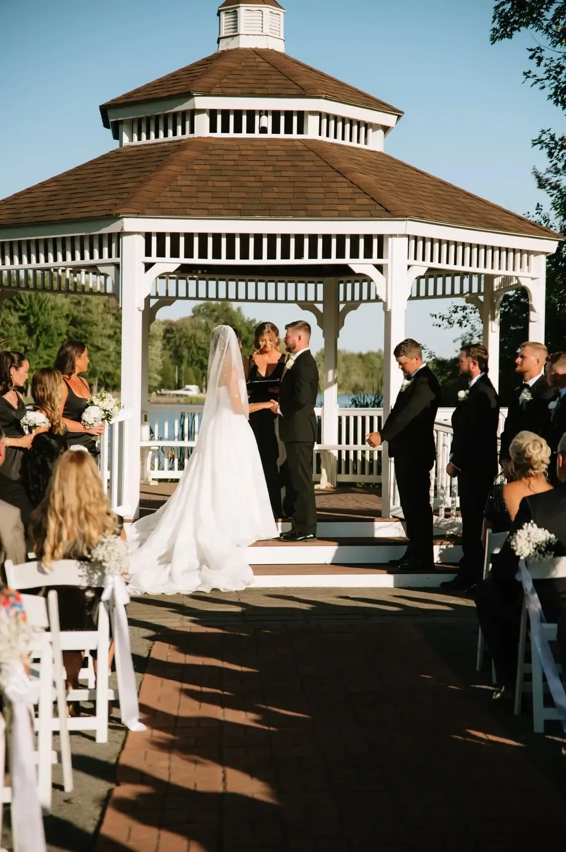 Wedding ceremony under a gazebo. Bride in white dress, groom in black suit, facing officiant. Guests seated.