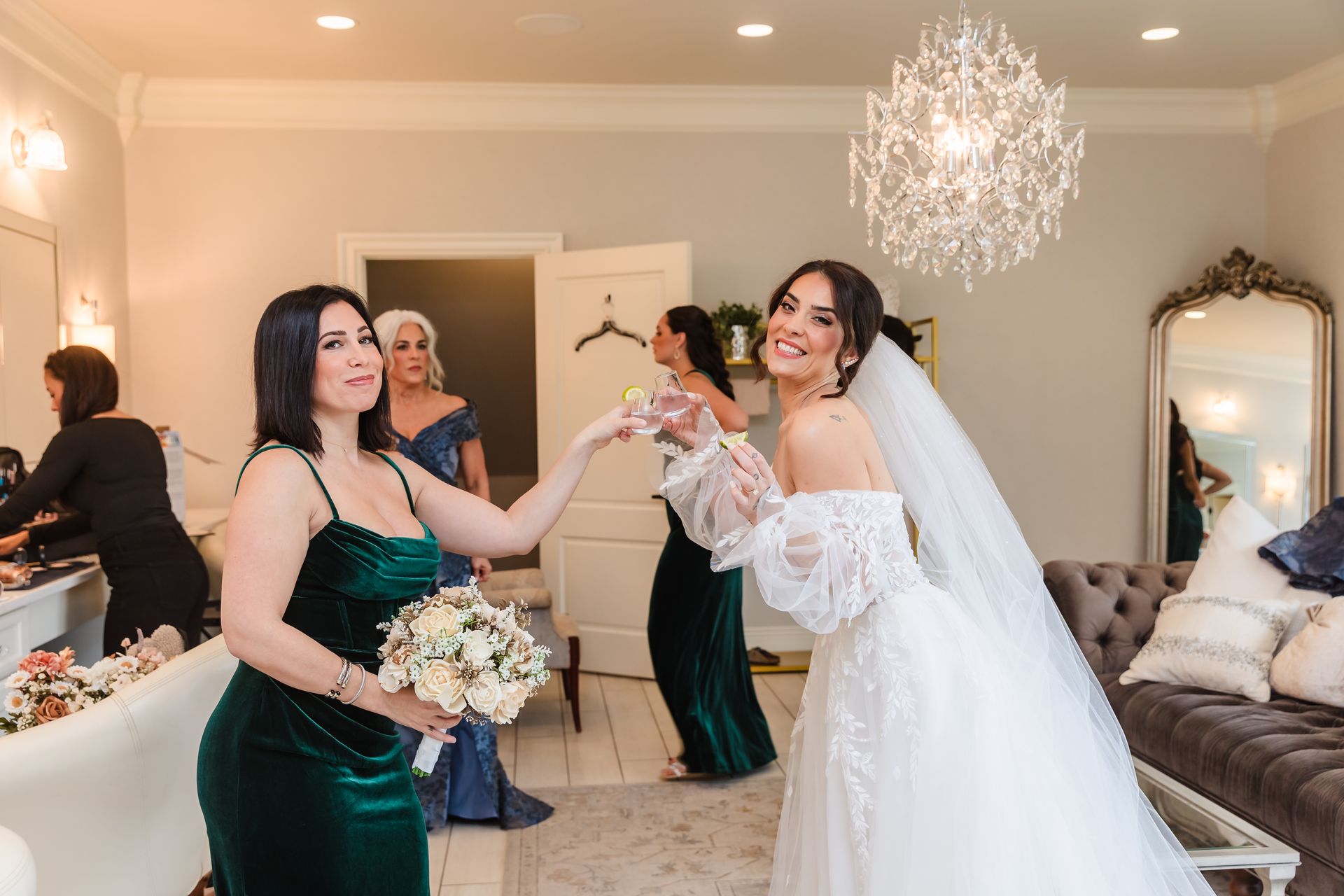 Bride in white dress celebrates with bridesmaid in green velvet dress, wedding prep room.