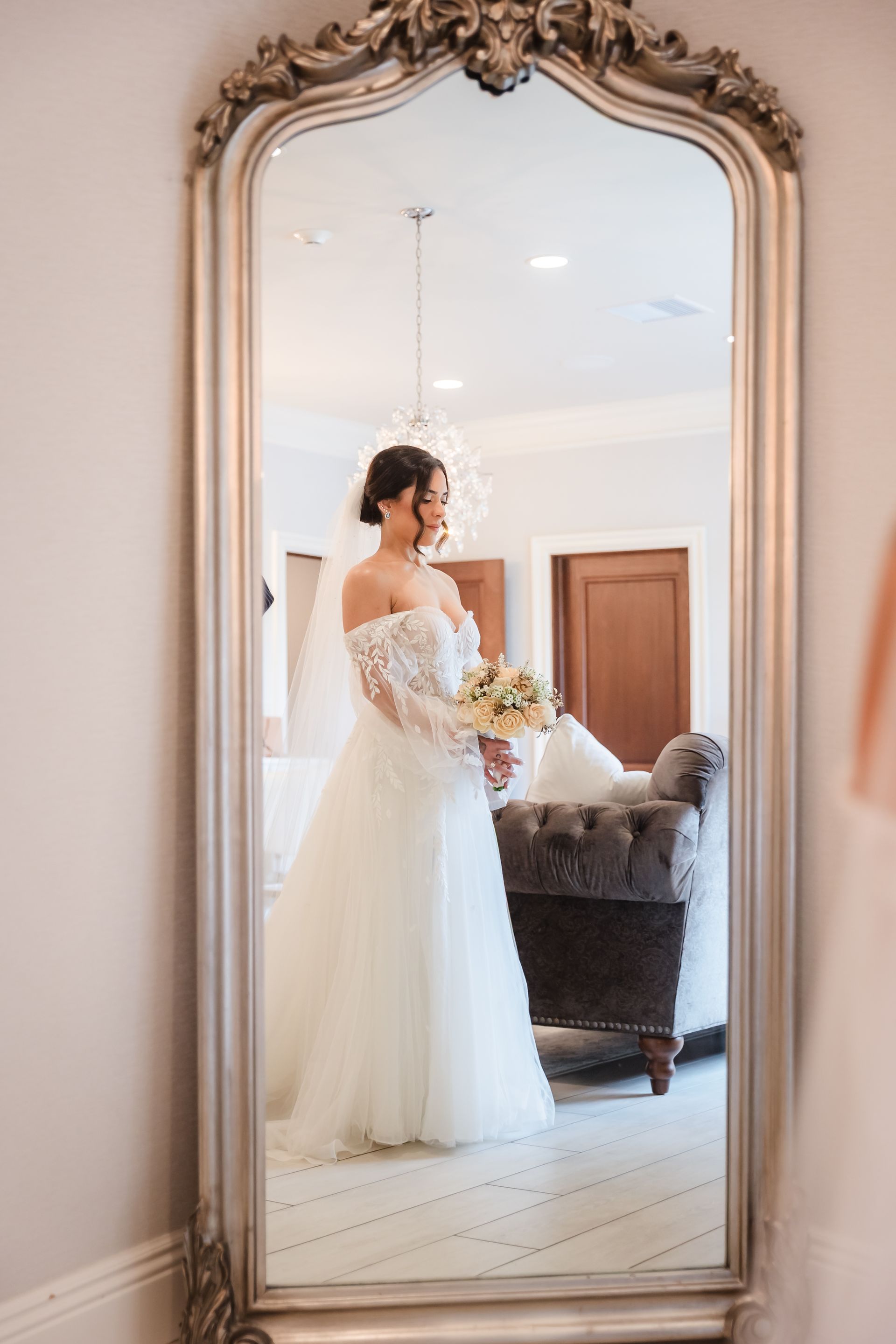 Bride in a white gown and veil smiles while holding flowers, reflected in a large ornate mirror.