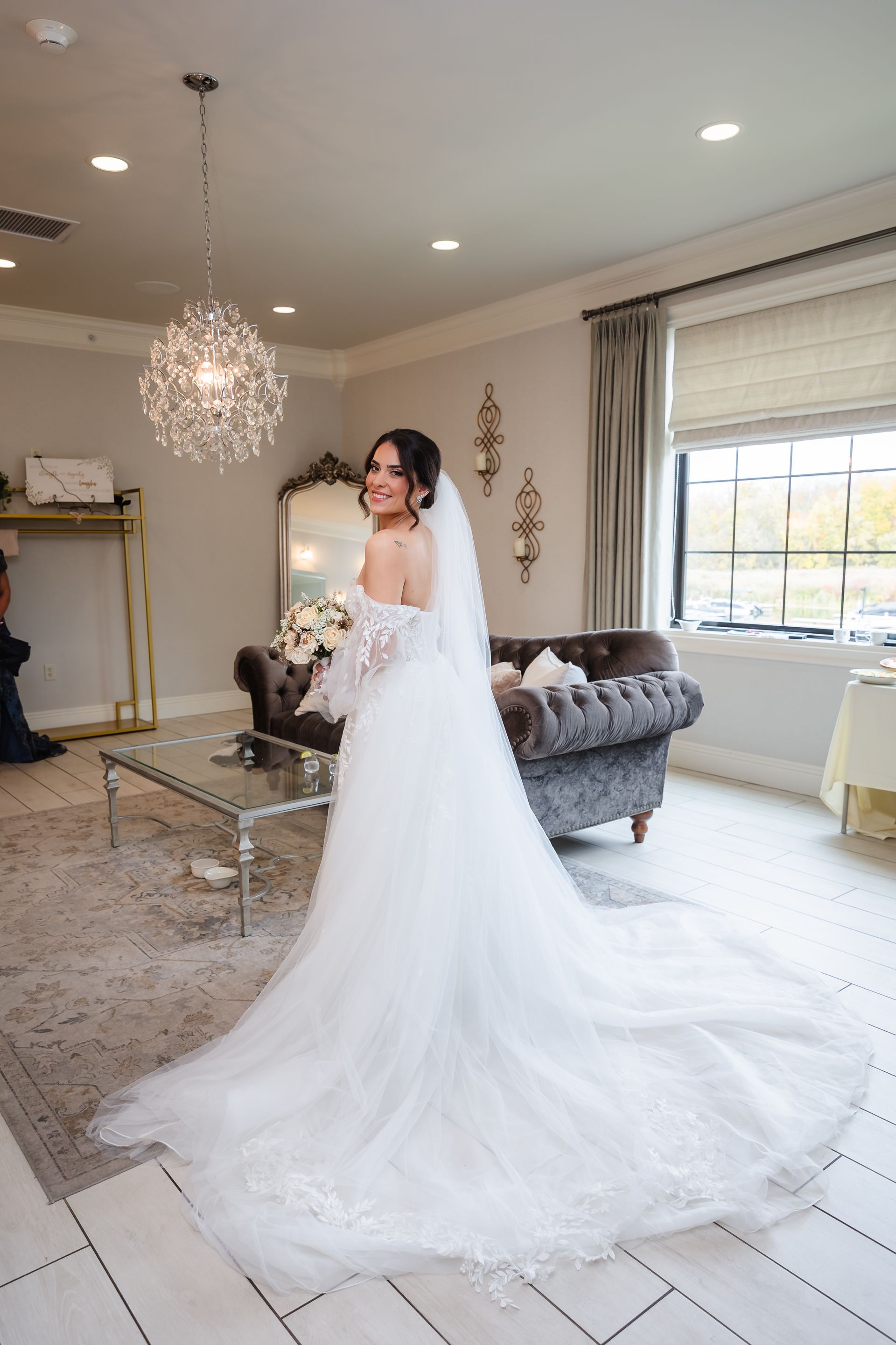 Bride in white off-shoulder gown, veil, smiling, looking over shoulder. Room with chandelier, couch, window.