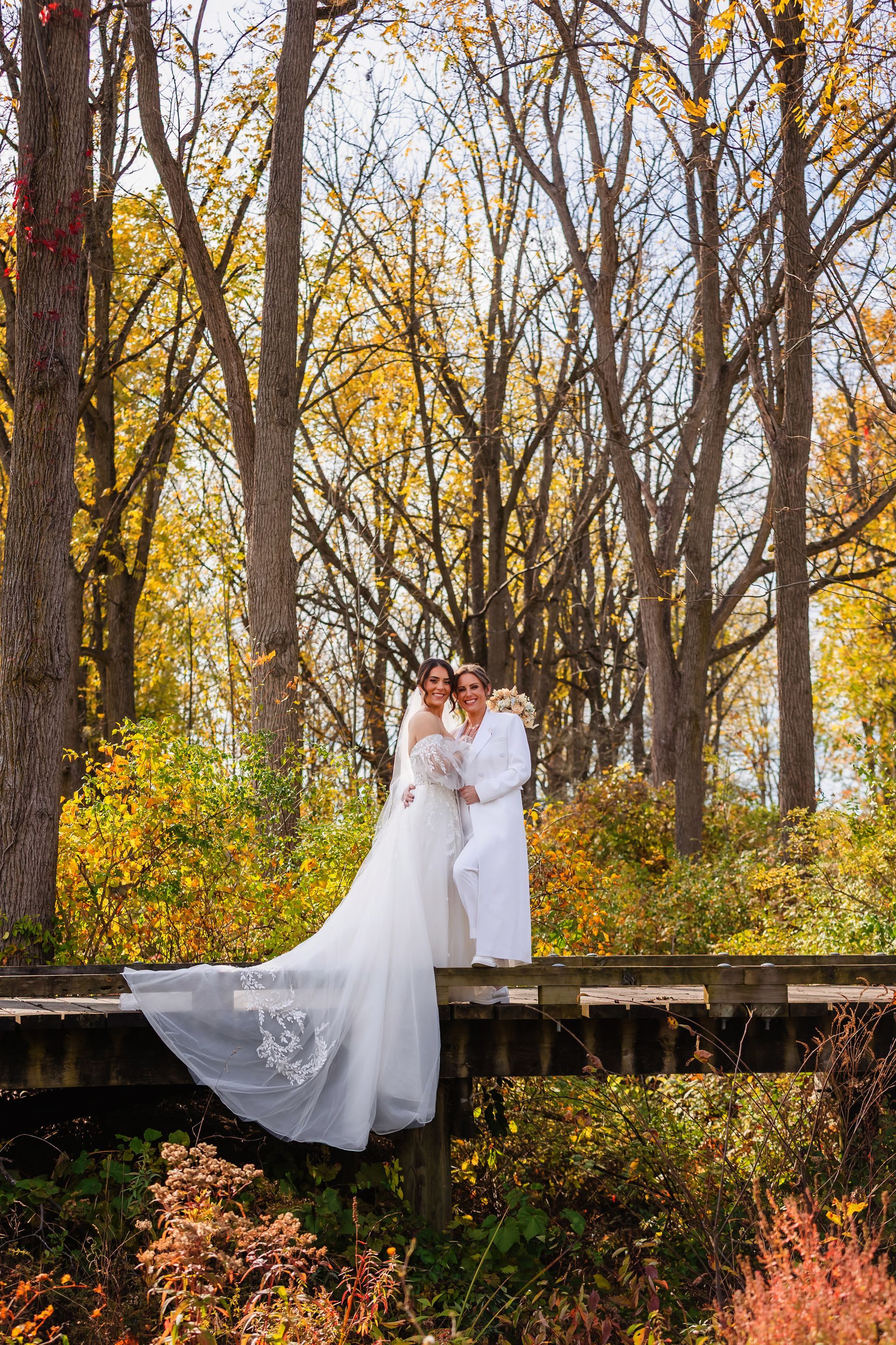 Couple in wedding attire on a wooden bridge, surrounded by autumn trees with yellow and orange leaves.