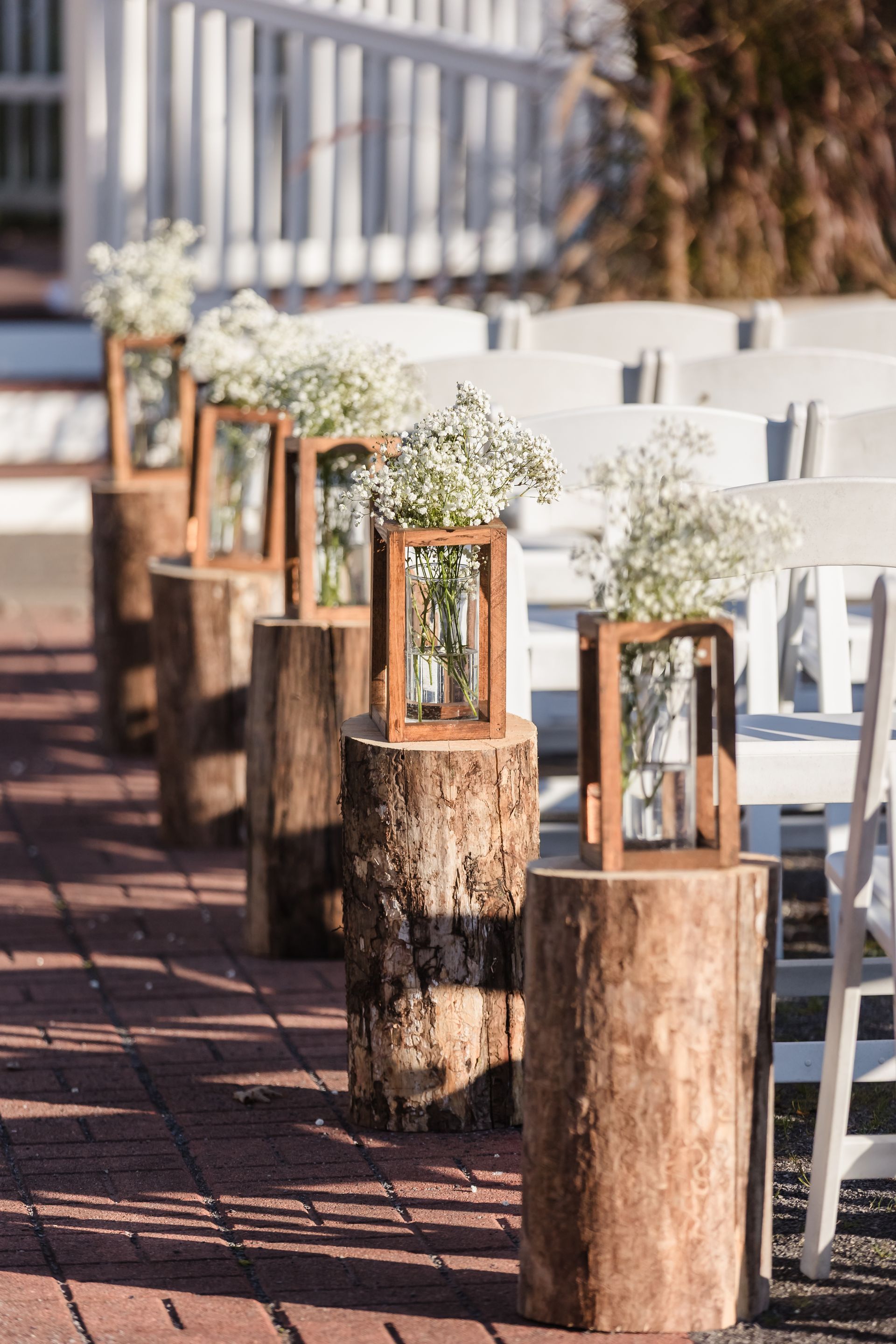 Wooden log pedestals with flower arrangements mark an outdoor wedding aisle, white chairs line the aisle.