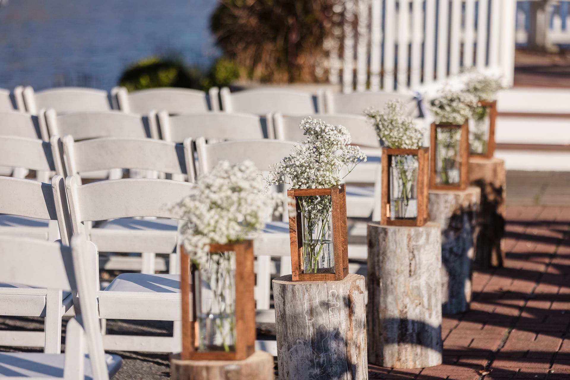 Rows of white chairs face wooden stumps with floral arrangements, waterside setting.