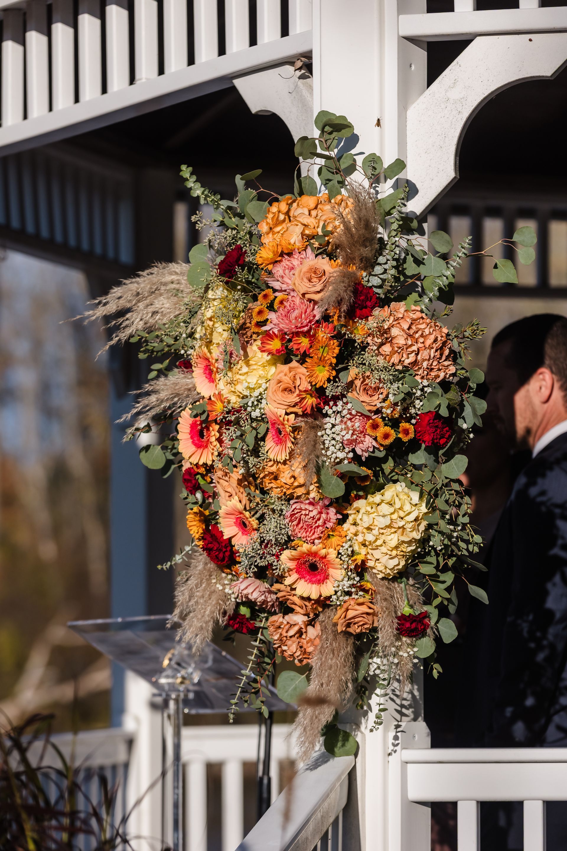 Floral arrangement on a white gazebo. Orange, pink, and red flowers with greenery. Man in suit to the right.
