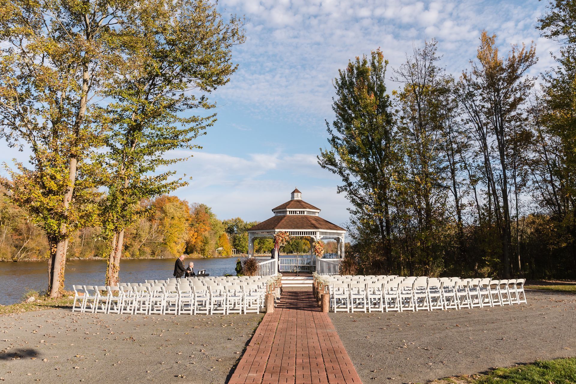 Wedding ceremony setting by a lake with a brick aisle, white chairs, and gazebo. Autumn trees and blue sky.