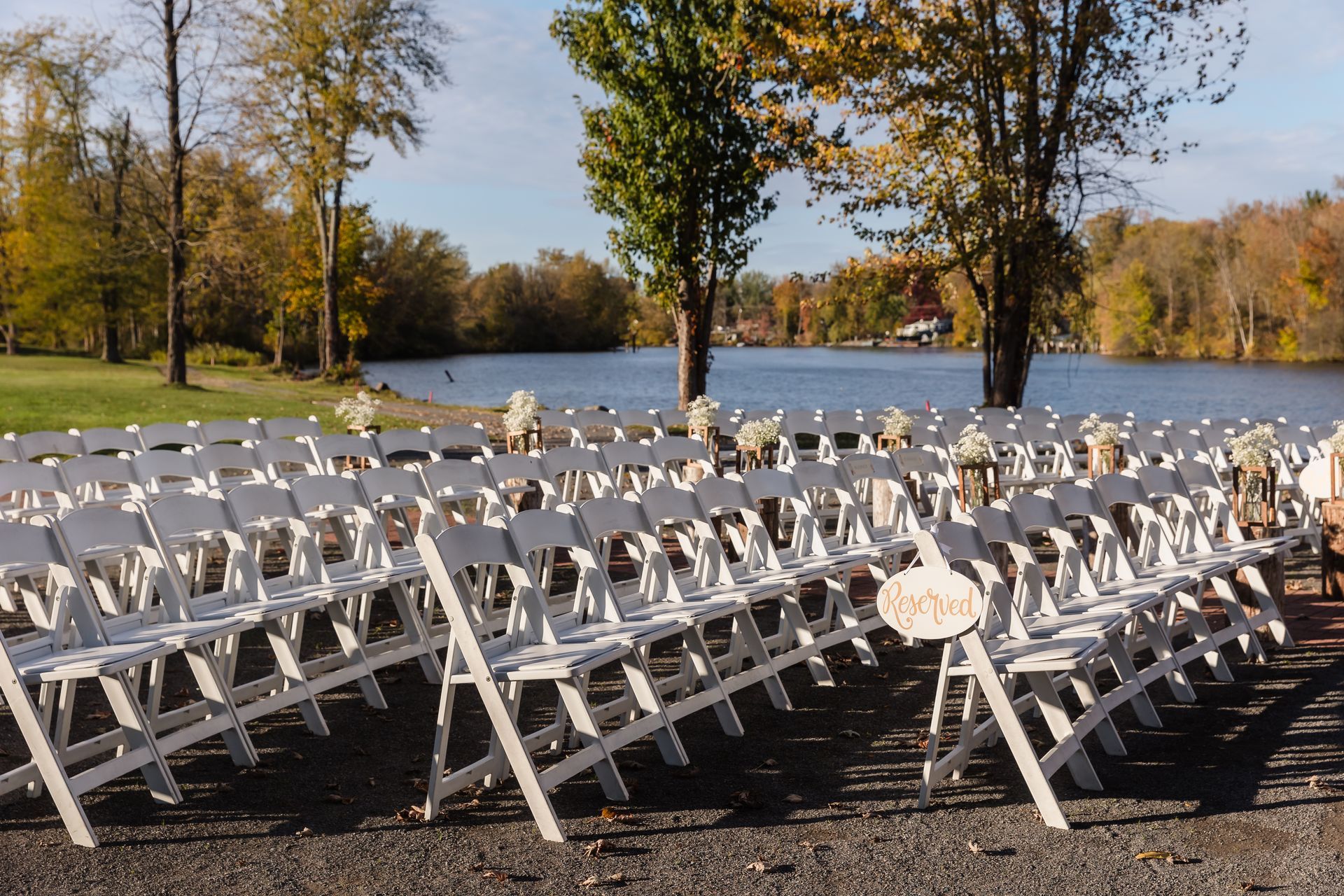 White chairs arranged for an outdoor wedding ceremony, with a lake and fall foliage in the background.