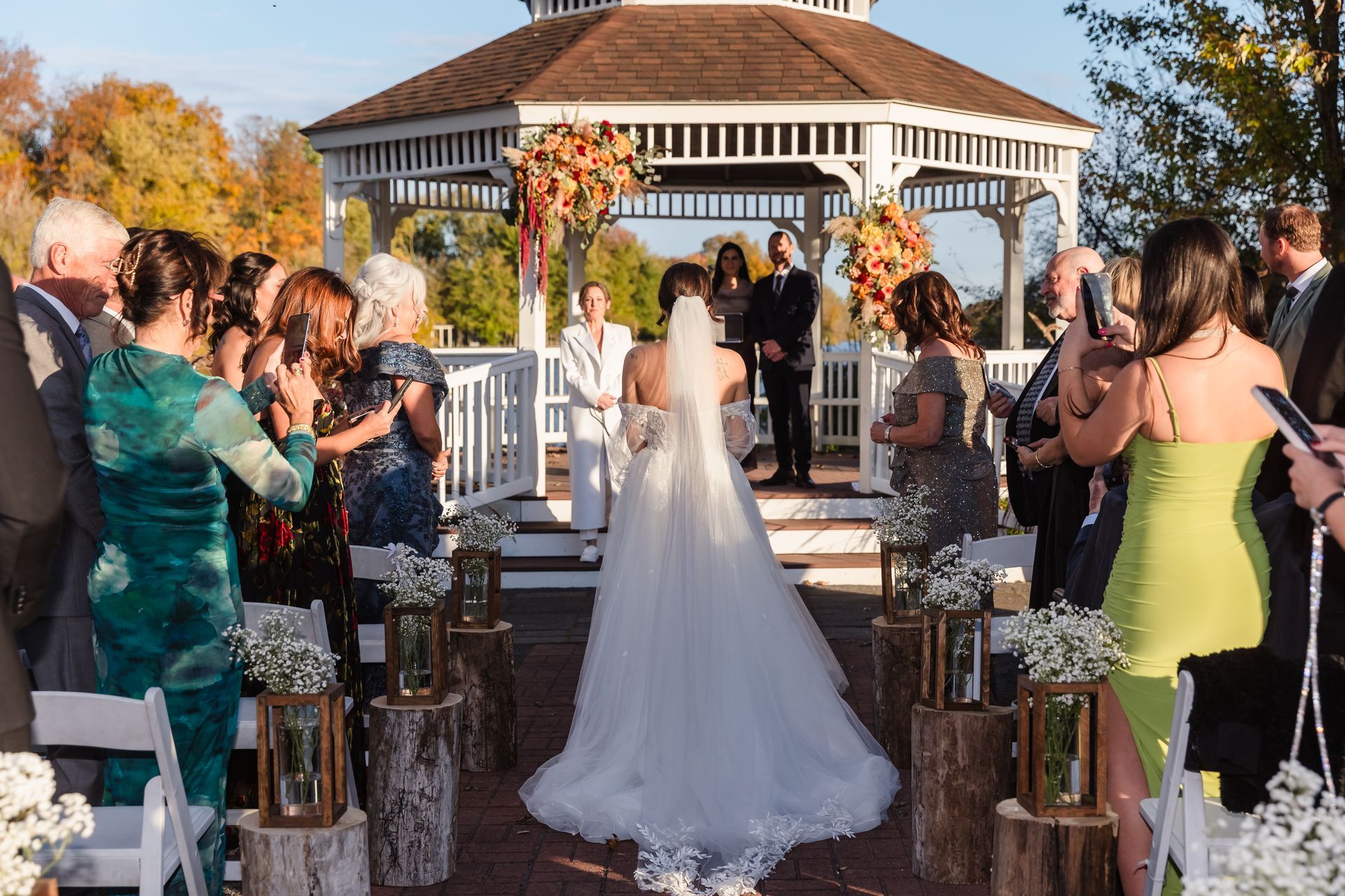 Bride walking down aisle to gazebo altar, guests taking photos. Outdoor wedding with fall foliage.