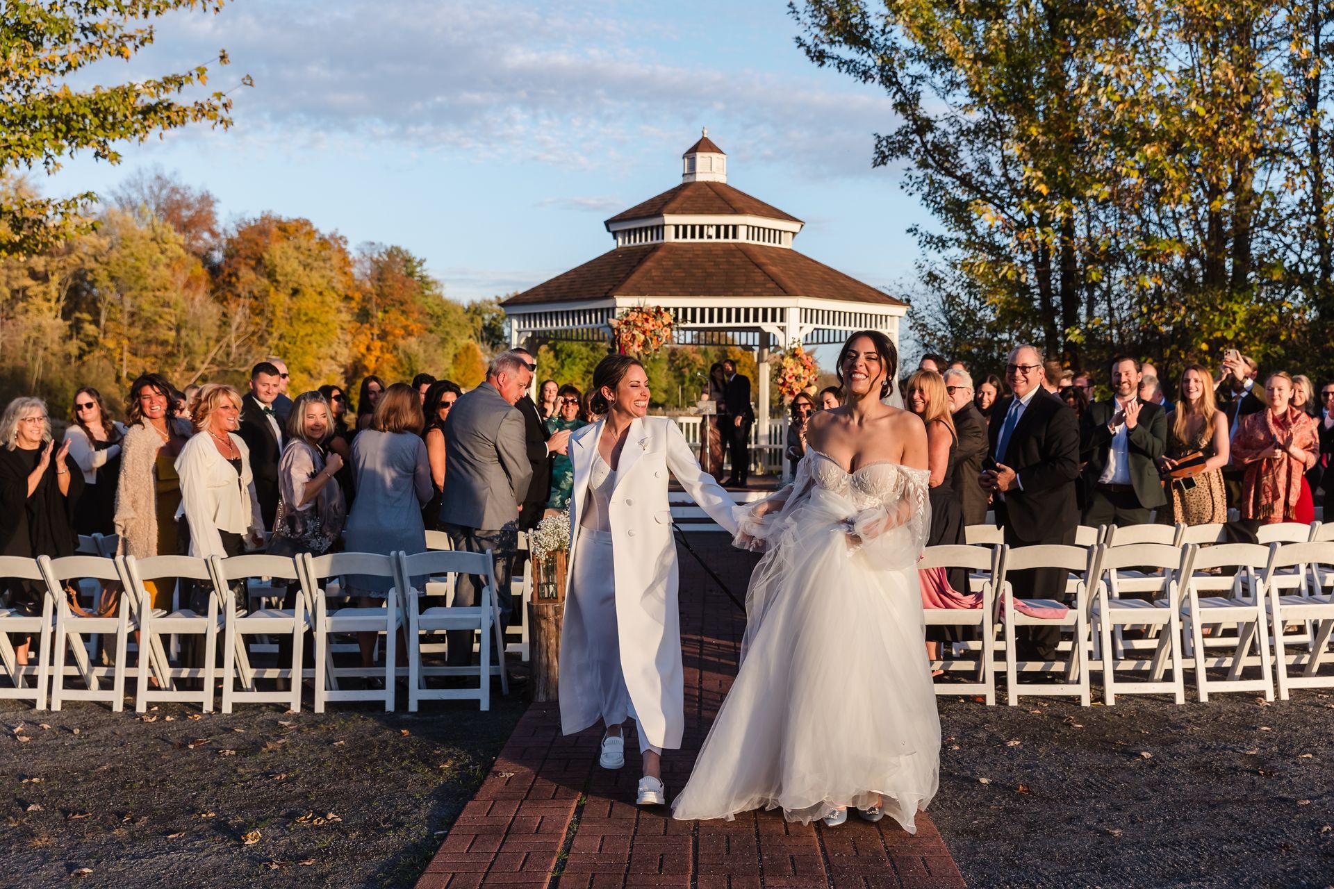 Two brides walking down a brick aisle after their wedding ceremony, guests cheering.