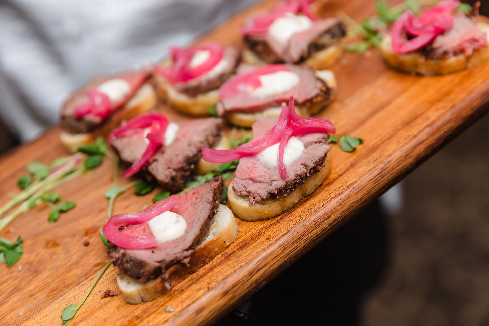 Wooden board of hors d'oeuvres: toast topped with meat, pink onions, and white sauce.