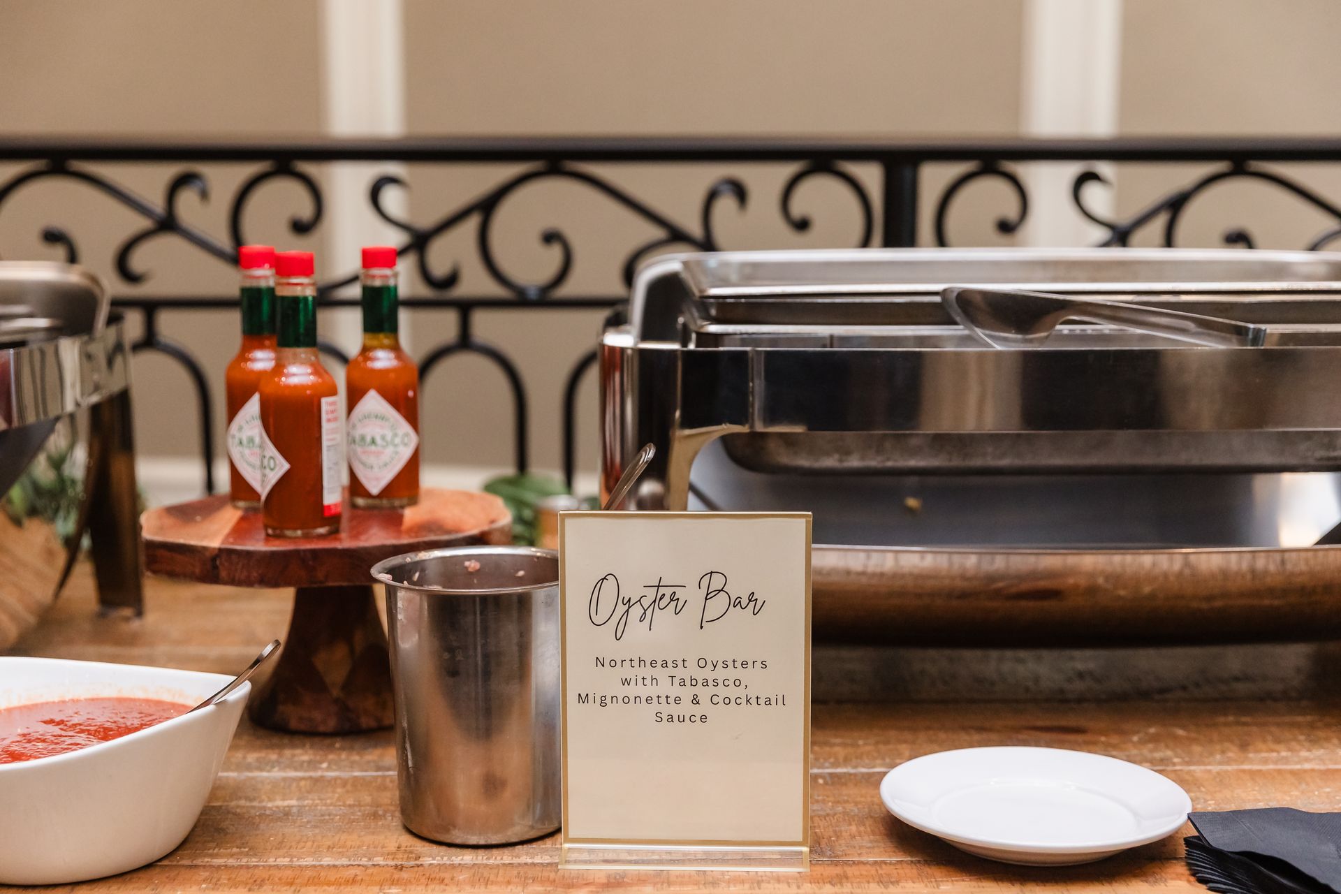 Buffet with hot sauce, chafing dish, food labels, and serving bowls on a wood table.