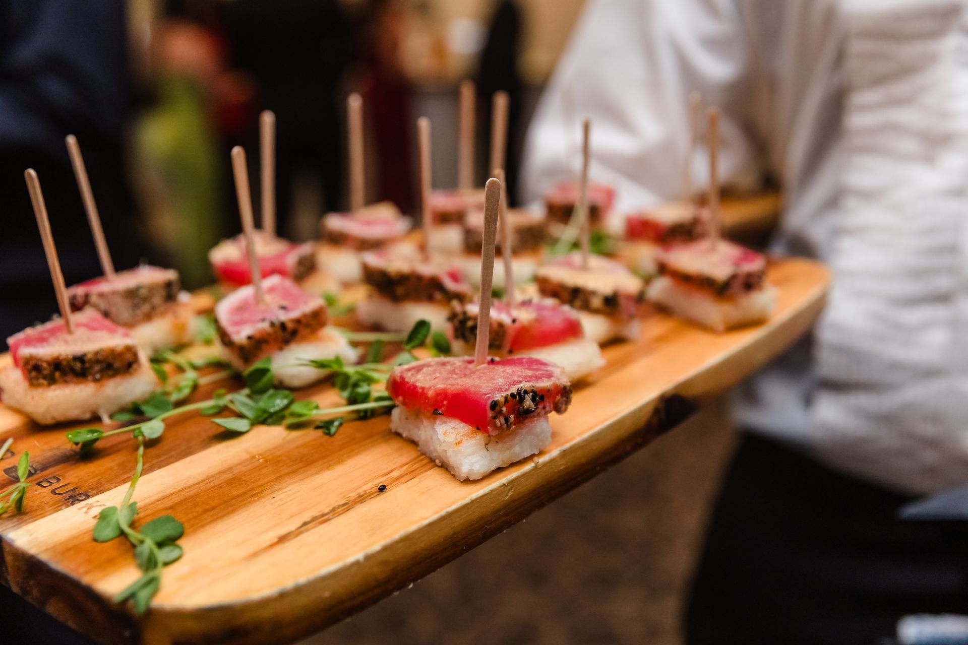 Wooden tray of tuna appetizers with toothpicks, held by person in white shirt.