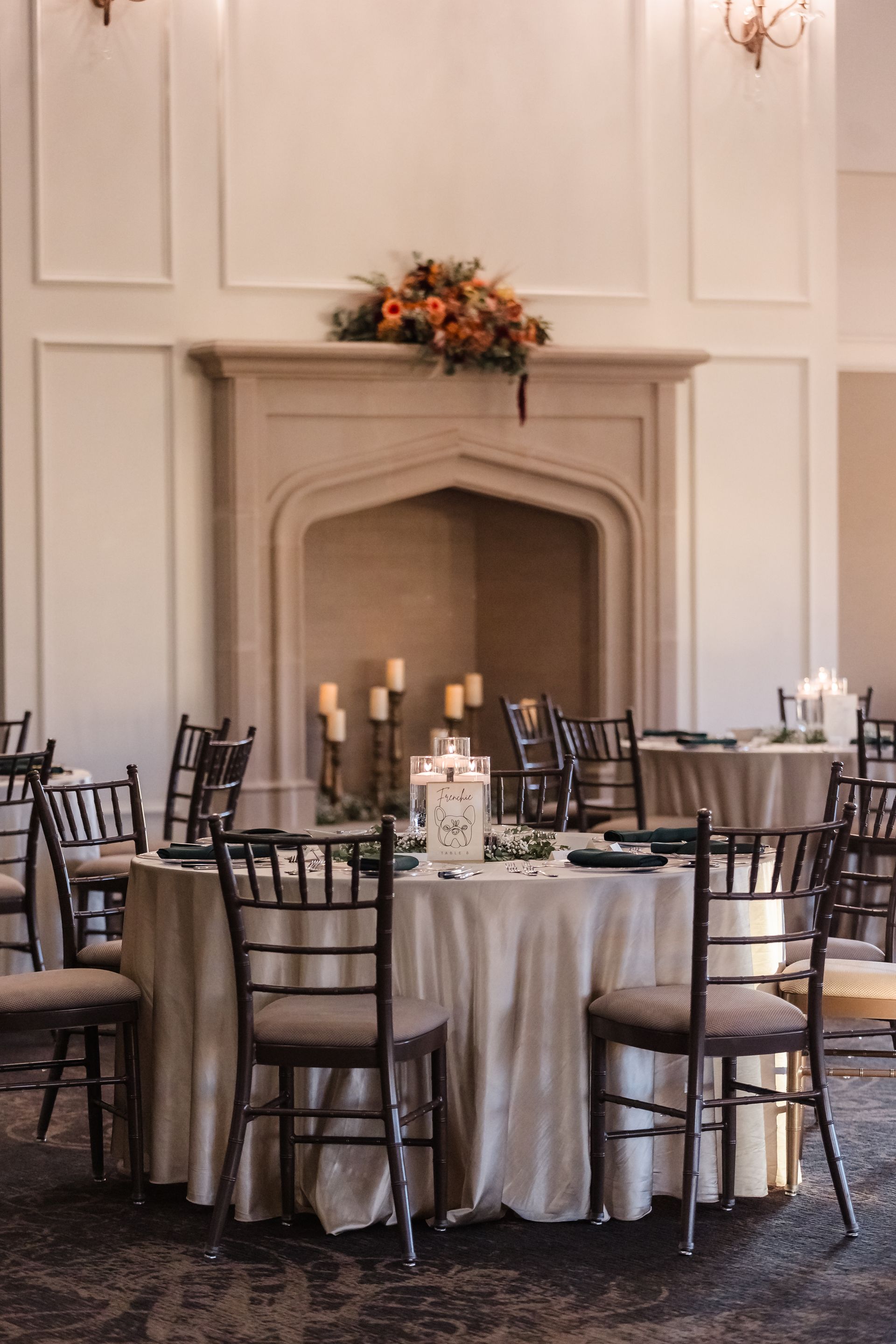 Wedding reception: round table with light cloth, dark chairs, fireplace with candles and floral arrangement.