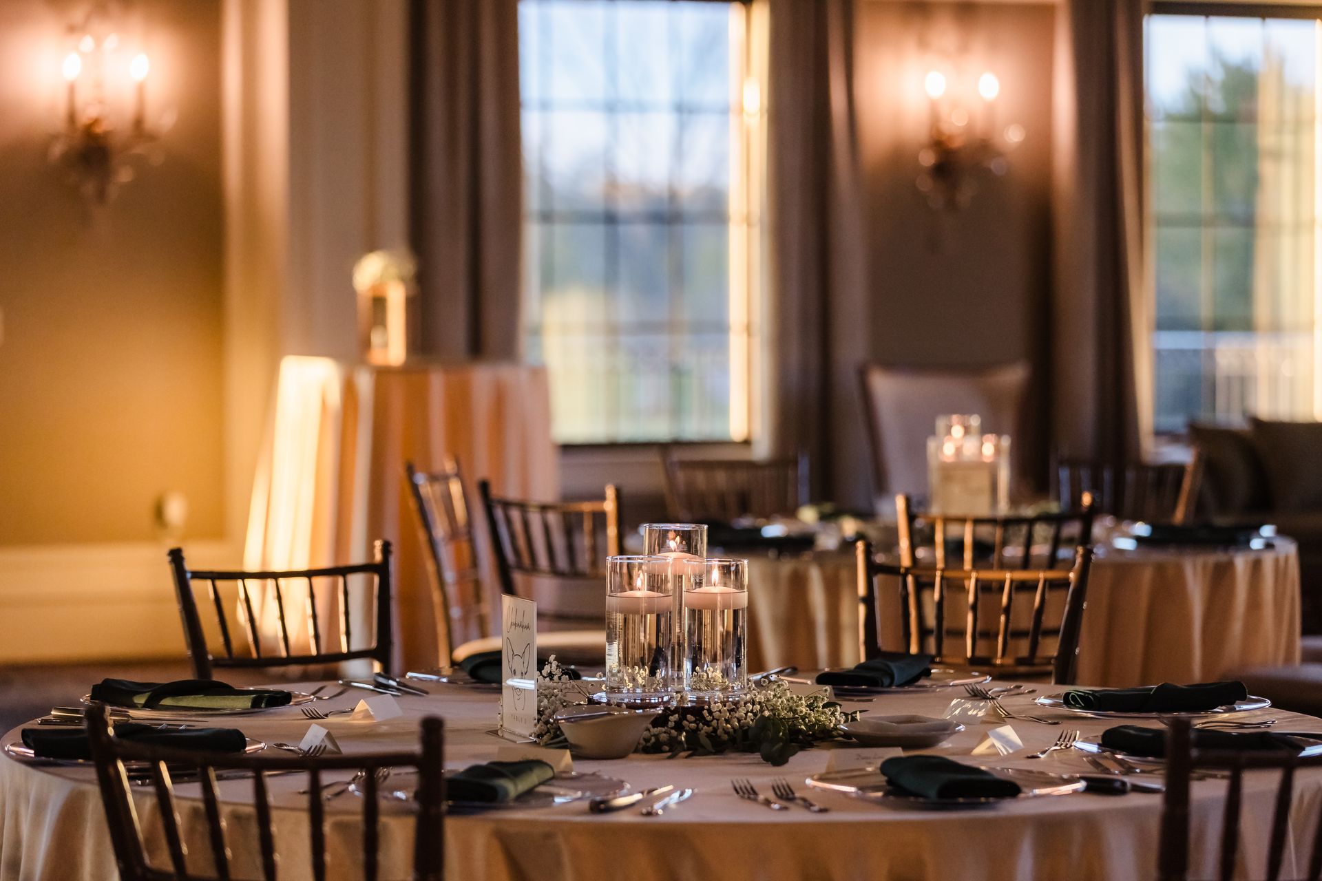 Elegant dining room set for a celebration. Round tables with beige linens, dark chairs, and centerpieces near a window.