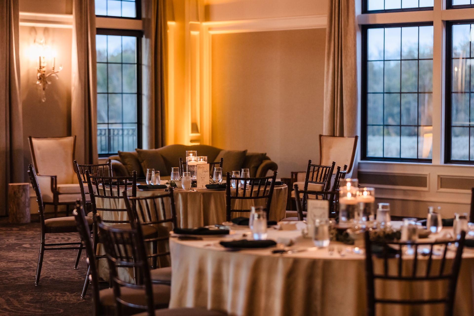 Elegant dining room set for an event; round tables with gold tablecloths, dark chairs, sofa, windows, and soft lighting.