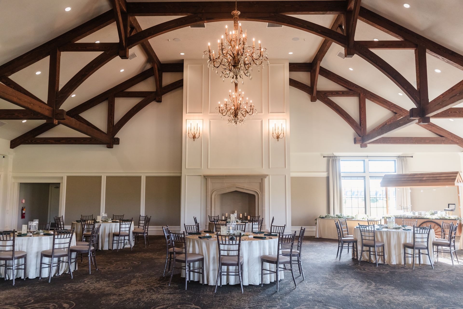 Elegant event space with round tables, chandeliers, and exposed wooden beams.