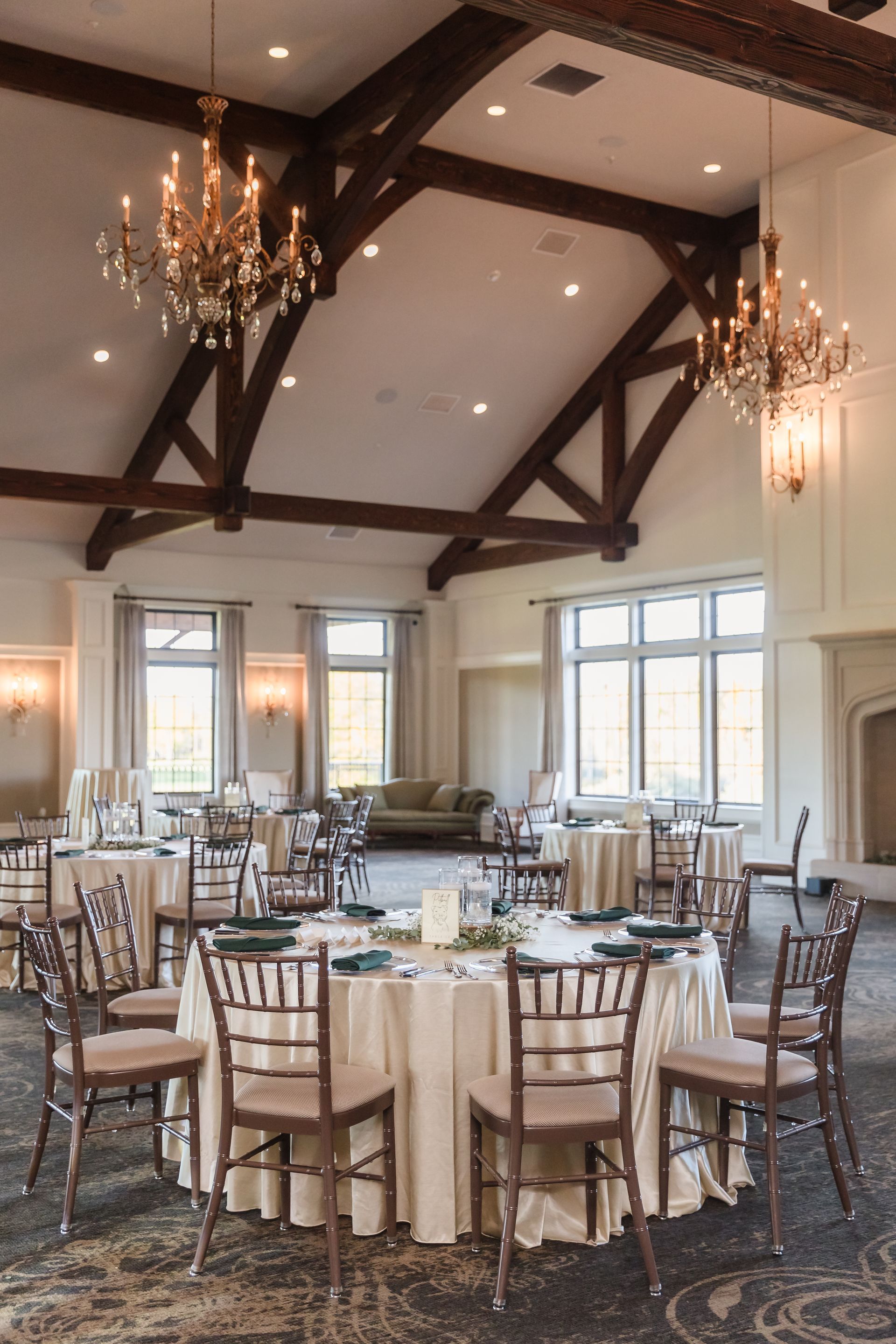 Elegant banquet hall with round tables, chairs, and chandeliers, beneath a high, beamed ceiling.
