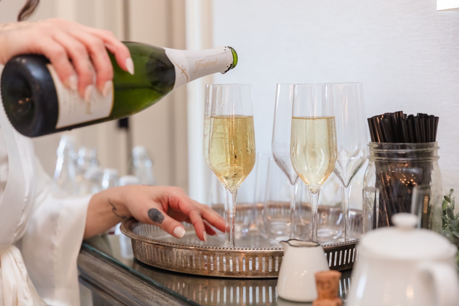 Woman pouring champagne from a green bottle into a glass, setting with a tray and a white pitcher.