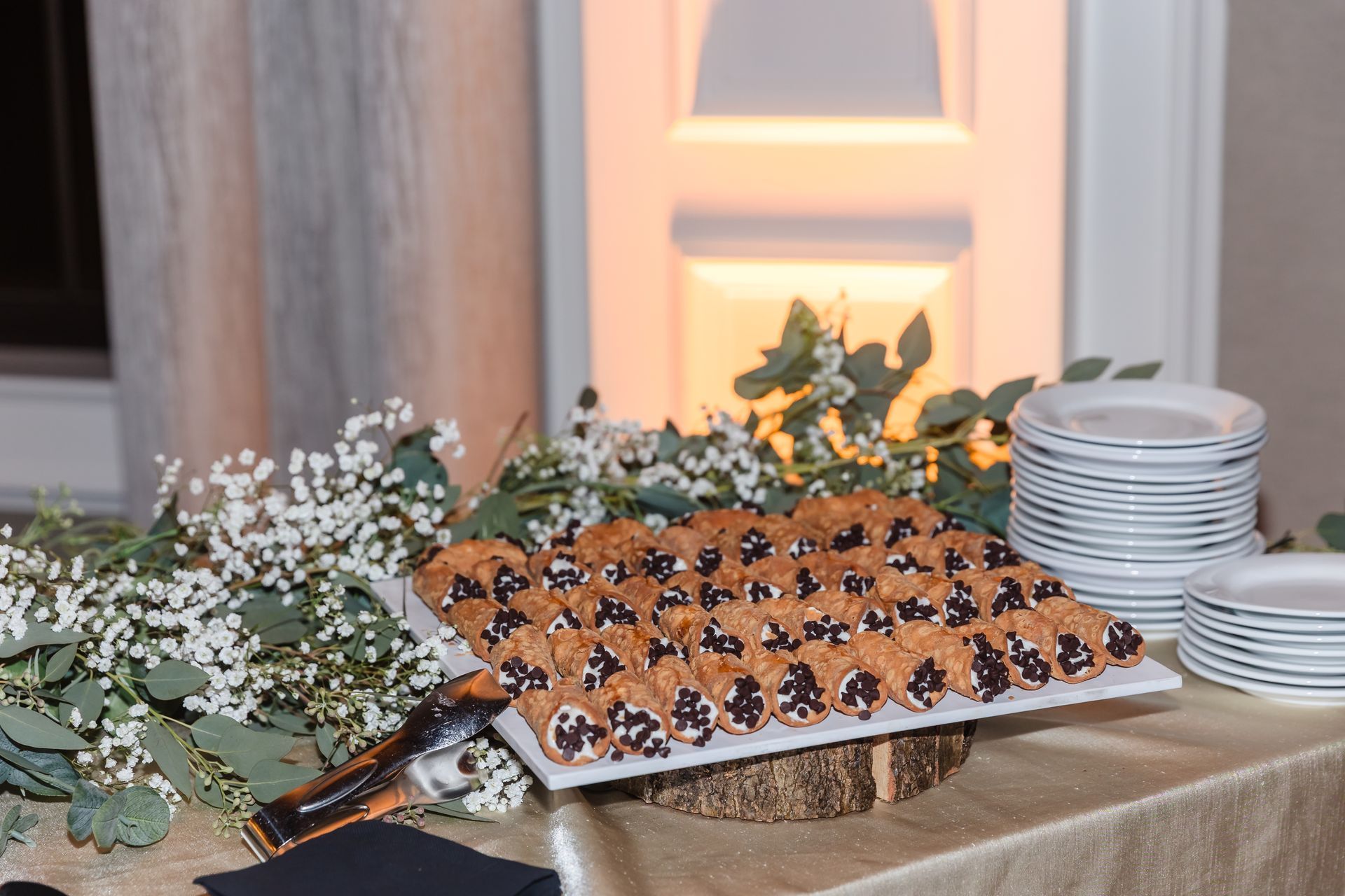 Cannolis on a platter, arranged on a wooden slice on a table with plates and greenery.