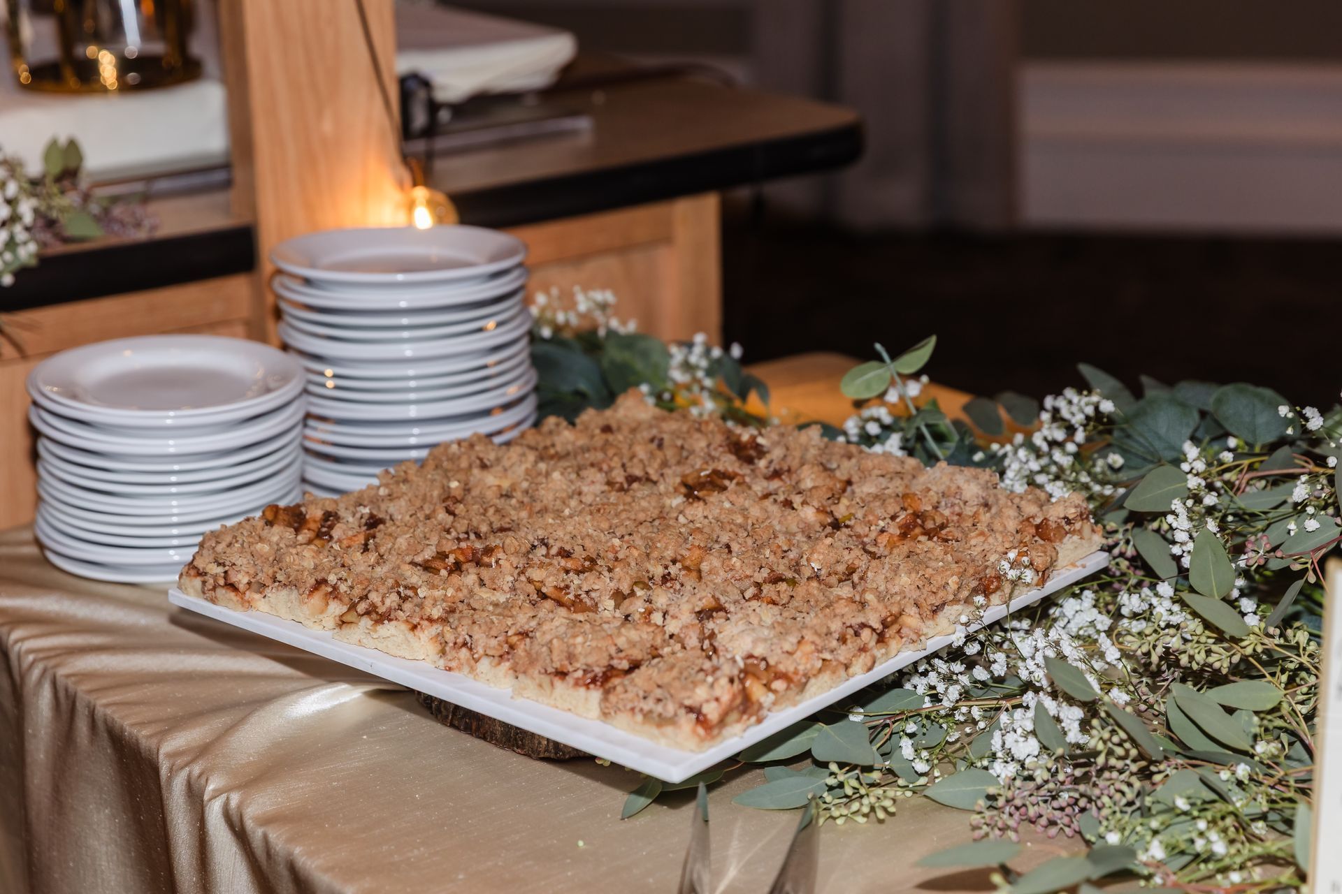 A dessert table features a square cake with crumb topping, white plates, and greenery on a gold tablecloth.