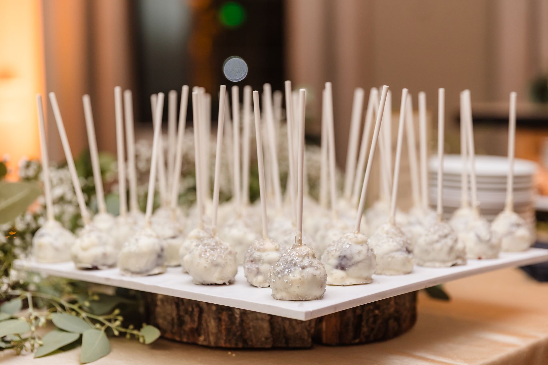Cake pops on a wooden base, covered in white frosting, on a white square platter.