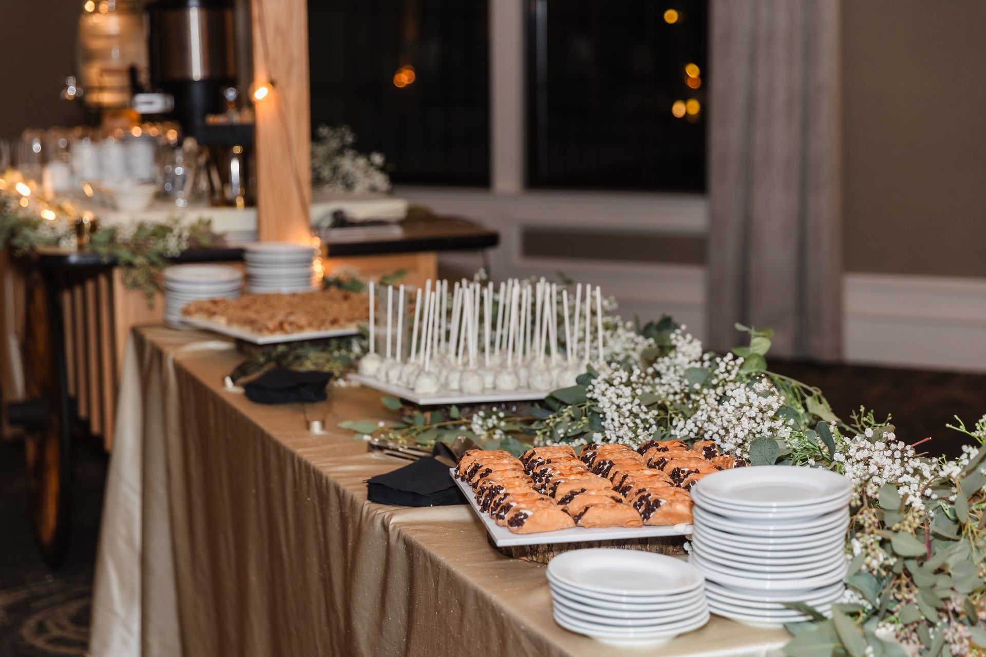 Dessert table with treats, stacked plates, and floral garland in a room with a window.
