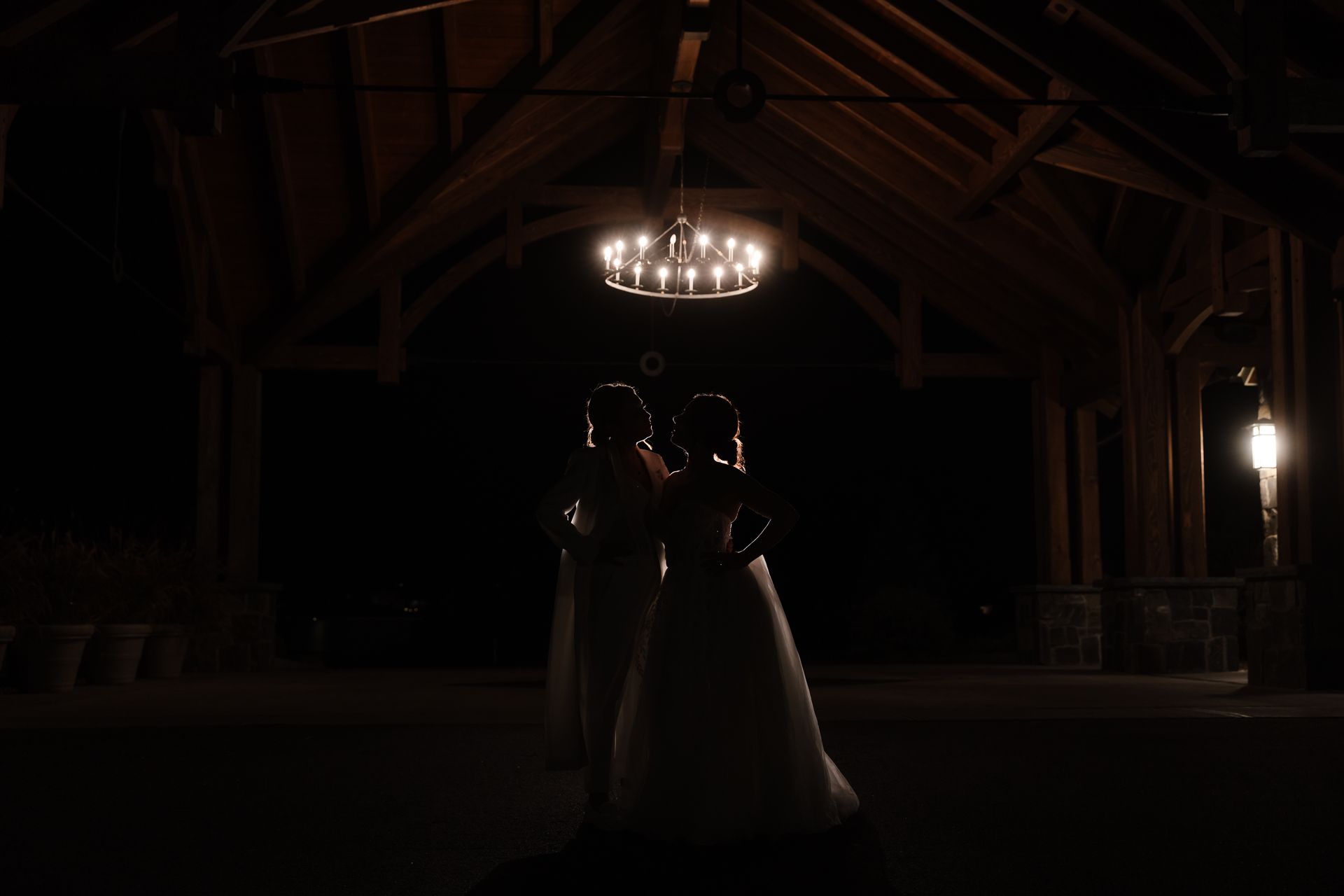 Two people in wedding gowns dance under a chandelier in a dimly lit wooden pavilion.