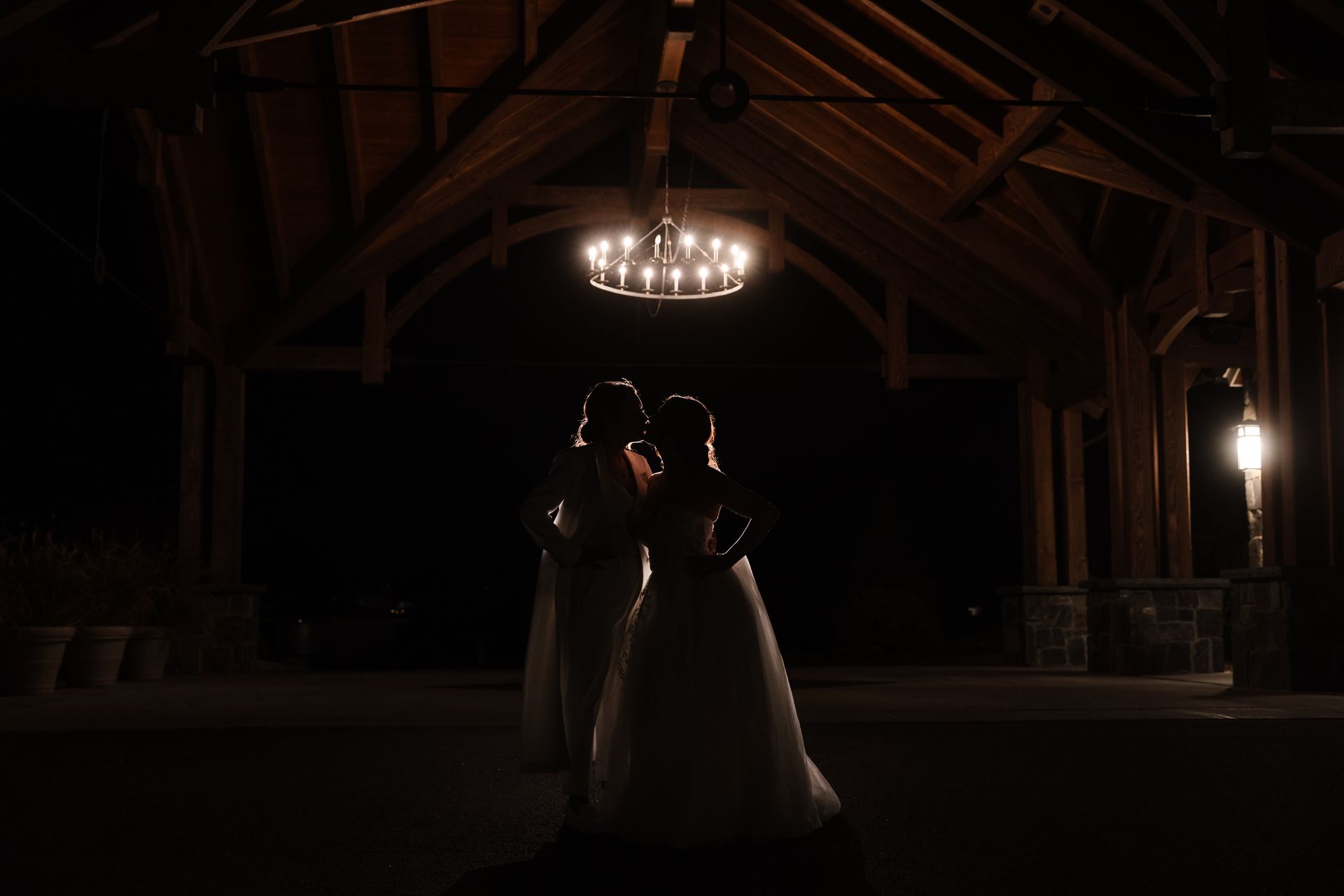 Two people in white gowns under a chandelier in a dark wooden structure.