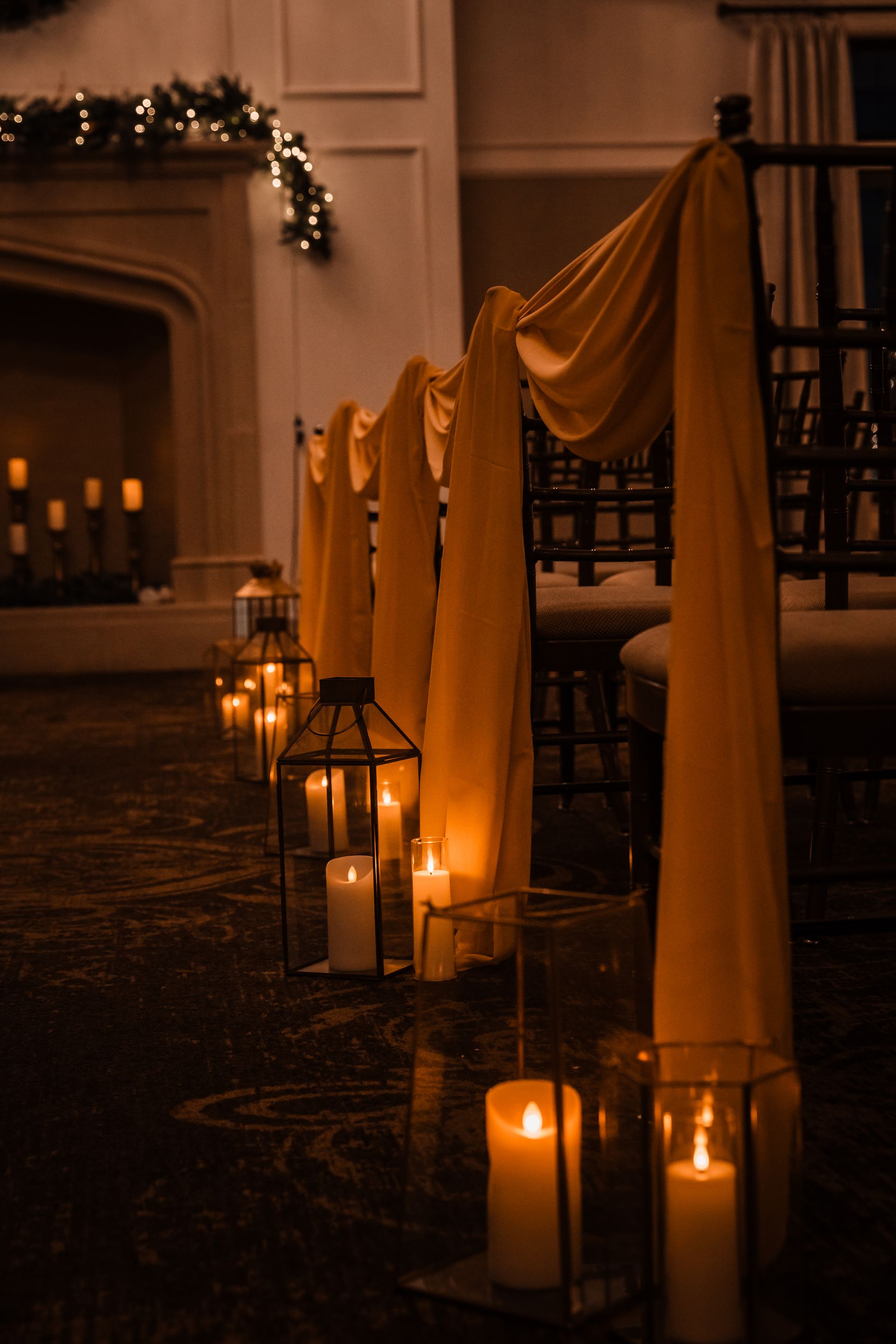 Candlelit ceremony aisle with draped chairs and lanterns near a fireplace decorated with lights.