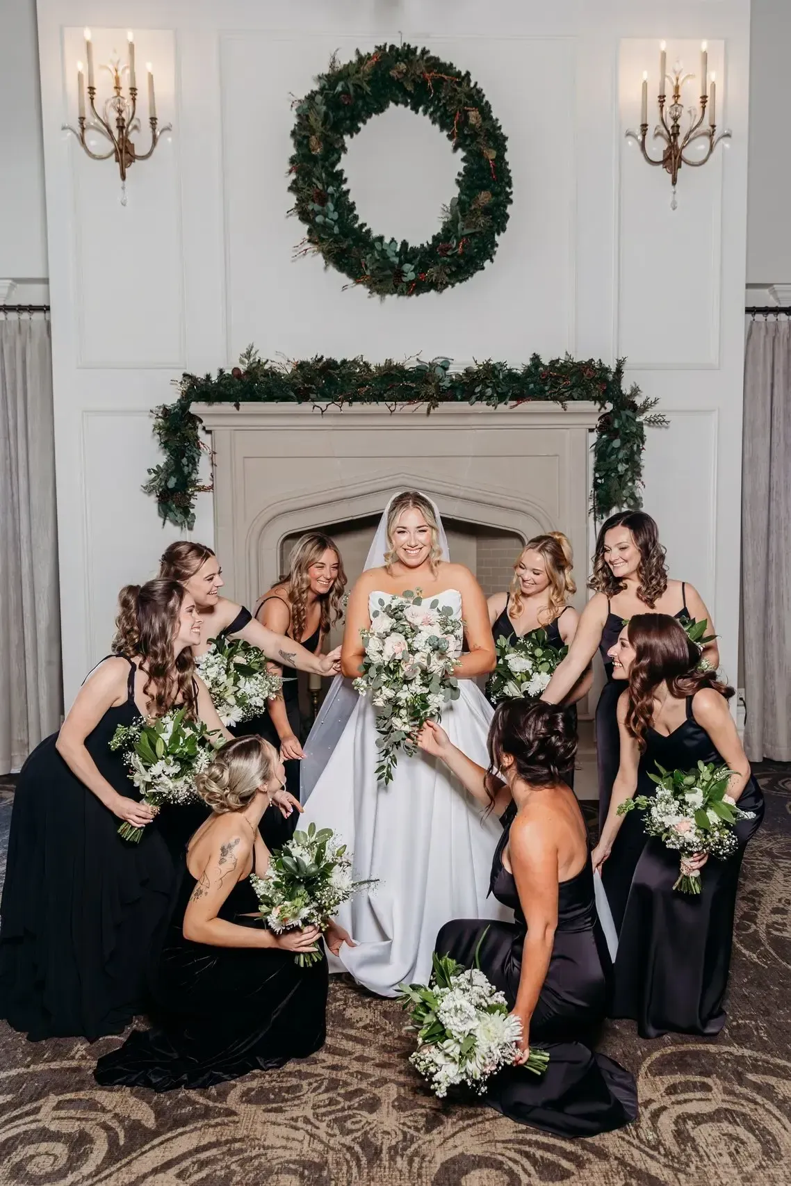 Bride in white gown surrounded by bridesmaids in black dresses, holding bouquets near a fireplace.