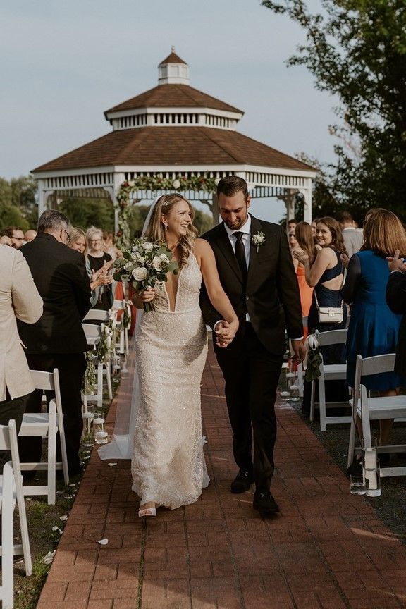 Newly married couple walking down aisle, smiling. Gazebo and guests in background.