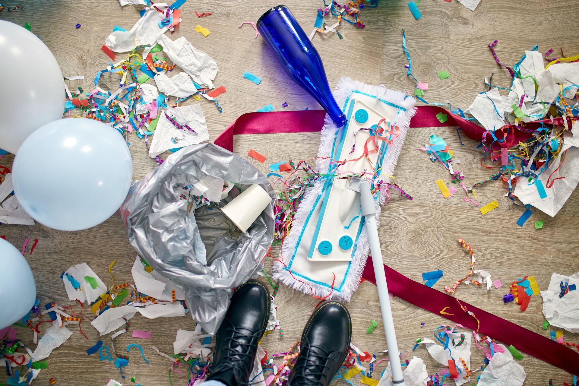 A person is standing on a messy floor with balloons , confetti and a mop.