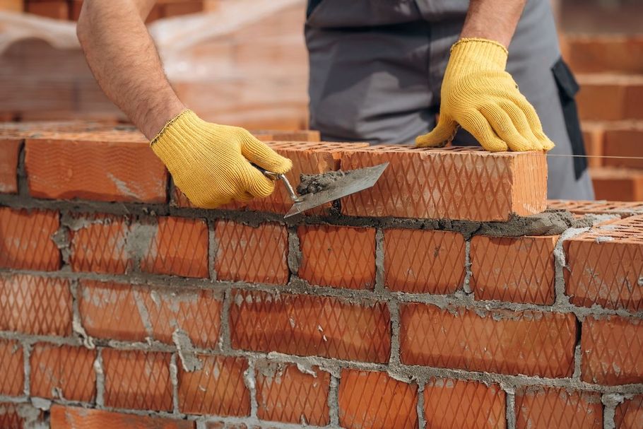 Worker laying red bricks with a trowel and yellow gloves at a construction site