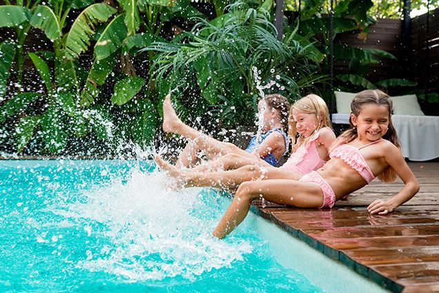 Three girls splashing in a pool, kicking their feet in the water. They wear swimwear and smile in a sunny setting.