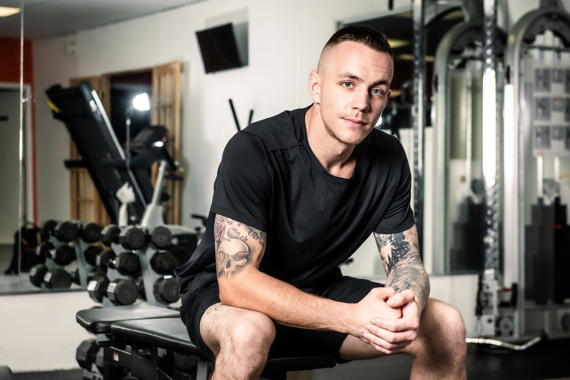 Man with tattoos sits on weight bench in a gym, looking at the camera.