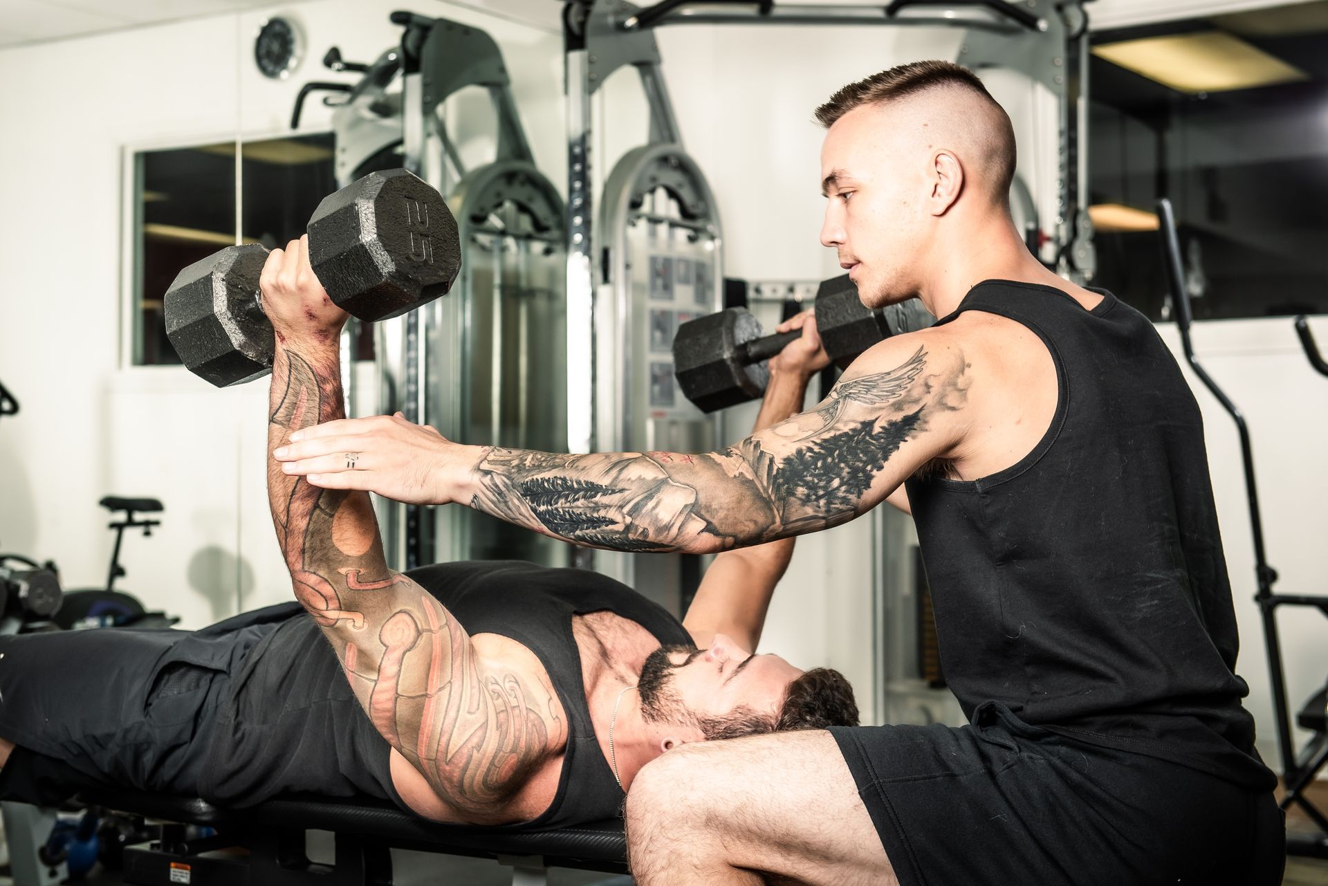 Man lifting dumbbells on a bench press, assisted by another man in a gym.