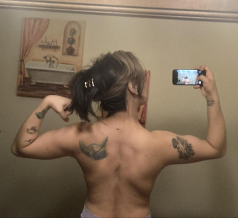 Person flexing back muscles, tattoos visible on arms and back. Holding phone, standing in front of a mirror.