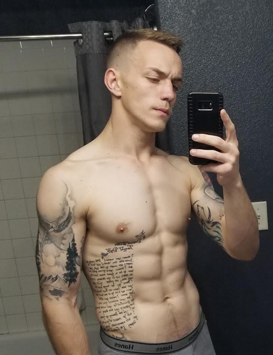 Man with toned physique takes a mirror selfie; he is shirtless with arm and side tattoos in a bathroom.