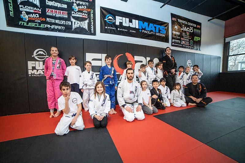 Group of martial arts students and instructors posing on red-and-black mats in a gym with FUJI MATS signage