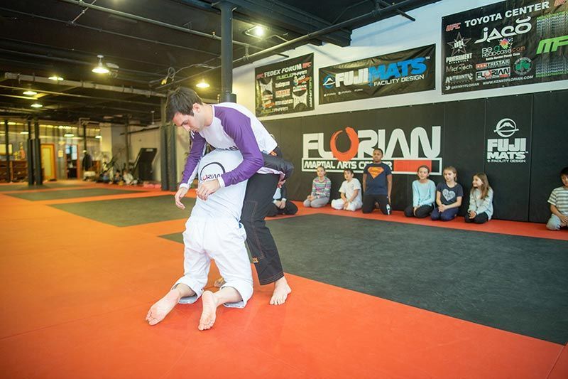 Two grapplers on an orange mat in a Jiu-Jitsu gym, one lifting the other as spectators watch.