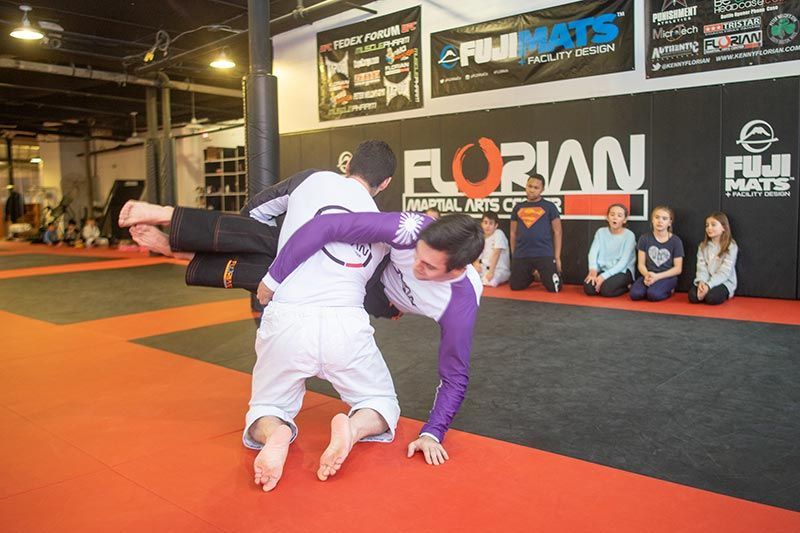Two jiu-jitsu fighters grapple on orange mats in a gym, with spectators seated behind them.