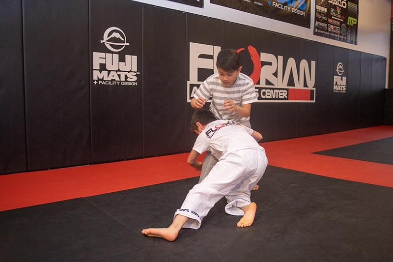Two martial artists practicing grappling on red-and-black mats in a gym with sponsor banners.